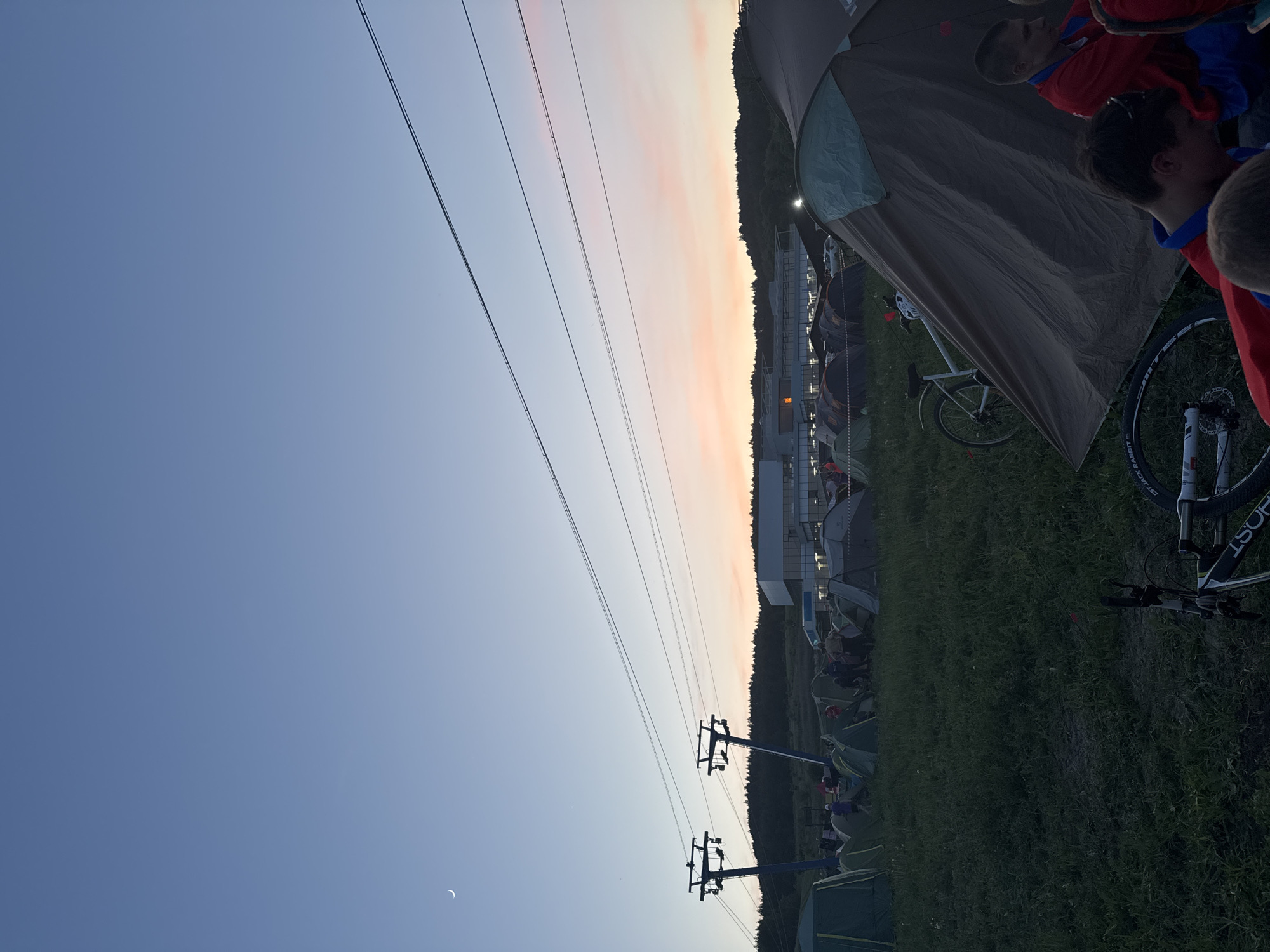 The image shows a campsite in Khvalynsk, Russia, during twilight. Visible are tents, people, and bicycles. A power line stretches across the sky, with support towers in view. The sky transitions from blue to a peach hue near the horizon. A crescent moon is faintly visible in the lower left corner of the sky. In the background, a building with lights on can be seen. The campsite is on grassy terrain.