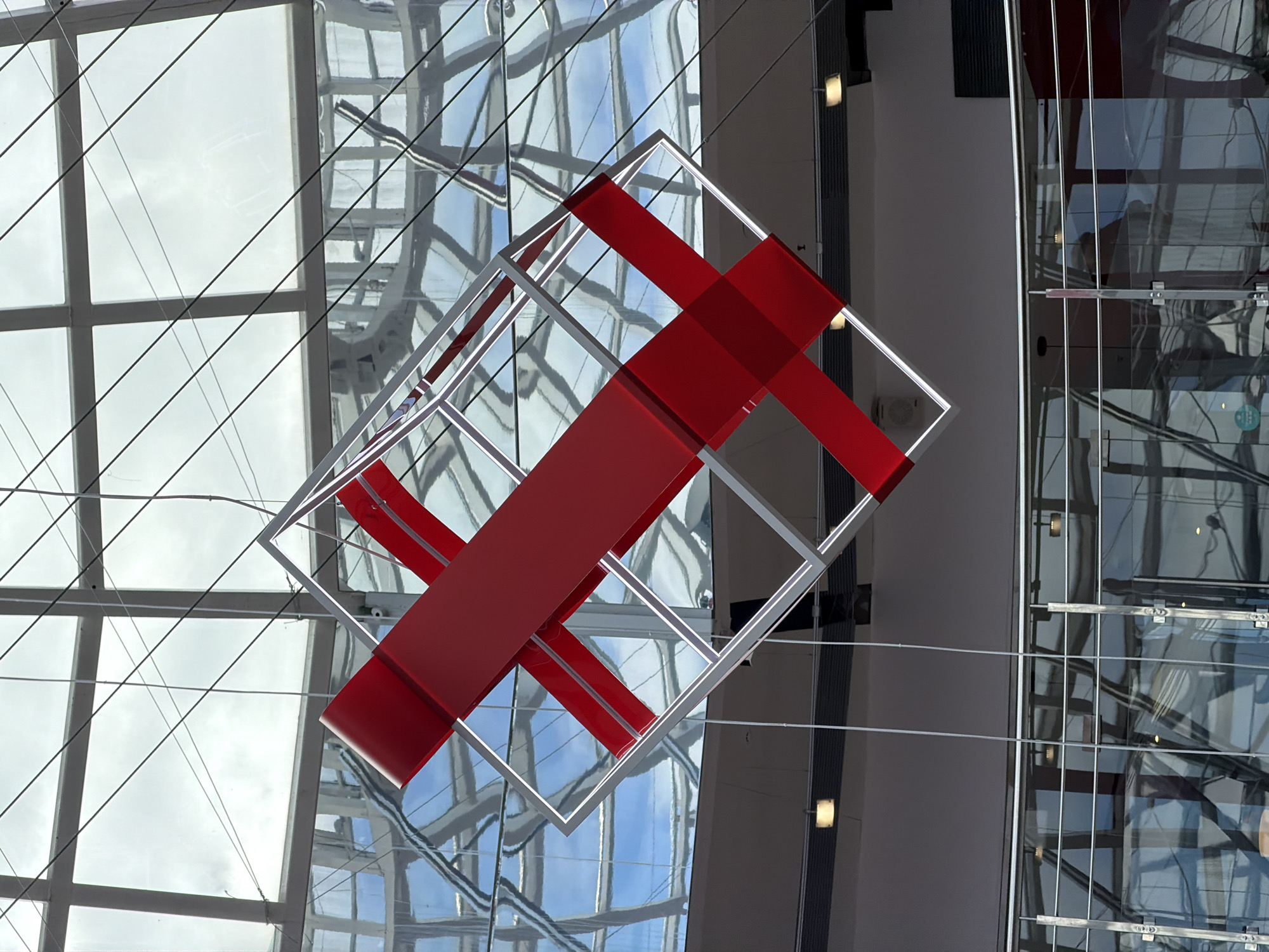 The image shows a modern art installation hanging in a large, bright space in Izhevsk, Russia. The installation consists of a white wireframe cube with two broad red bands intersecting within it. The cube hangs suspended by thin wires from a grid-like ceiling. The ceiling is comprised of numerous glass panels, reflecting the sky outside. In the background, there are architectural features including glass walls, floors, and supportive structures. Some of these also reflect parts of the sky, indicating the presence of a large atrium or similar structure. The general impression is of a modern, open, and airy building.