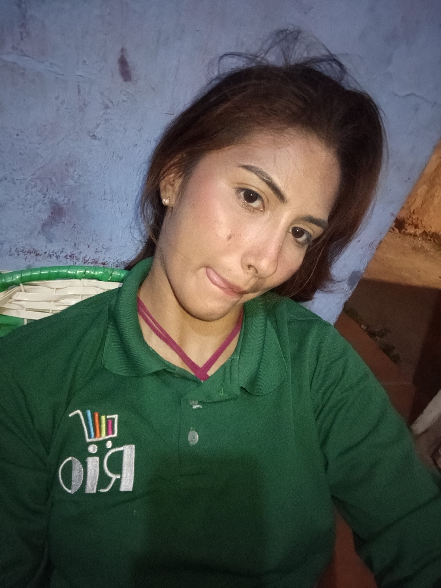 A close-up, medium-shot, selfie-style photograph shows a young woman with light brown hair and fair skin wearing a dark green polo shirt with a white embroidered logo. The woman has make-up on, including foundation, blush, and eye make-up. Her left eyebrow is arched and her mouth is slightly open with her tongue peeking out. She is wearing a thin, dark pink strap around her neck. The background is a textured, light blue wall. To the left of the woman, a portion of a green woven object is visible. The lighting in the image suggests it was taken indoors or at dusk, with a warm, yellowish light source illuminating the right side of the frame. The image is likely taken in Juan Griego, Venezuela.