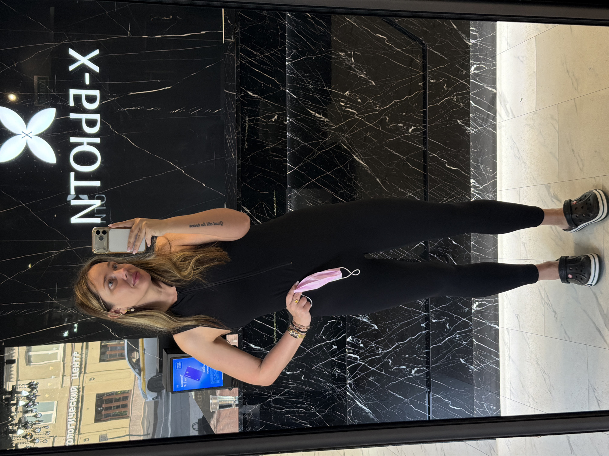 A woman stands in front of a mirrored wall, taking a selfie. She is wearing a black sleeveless jumpsuit and black Crocs. She is holding a pink face mask in her right hand. In the reflection of the mirror, we see a street scene with buildings and a white car. The wall behind her is black marble with white veining. A logo reading "NTOLPA-X" is visible on the wall. The floor is tiled in a light color. The image appears to be taken indoors, likely in a salon or shop.