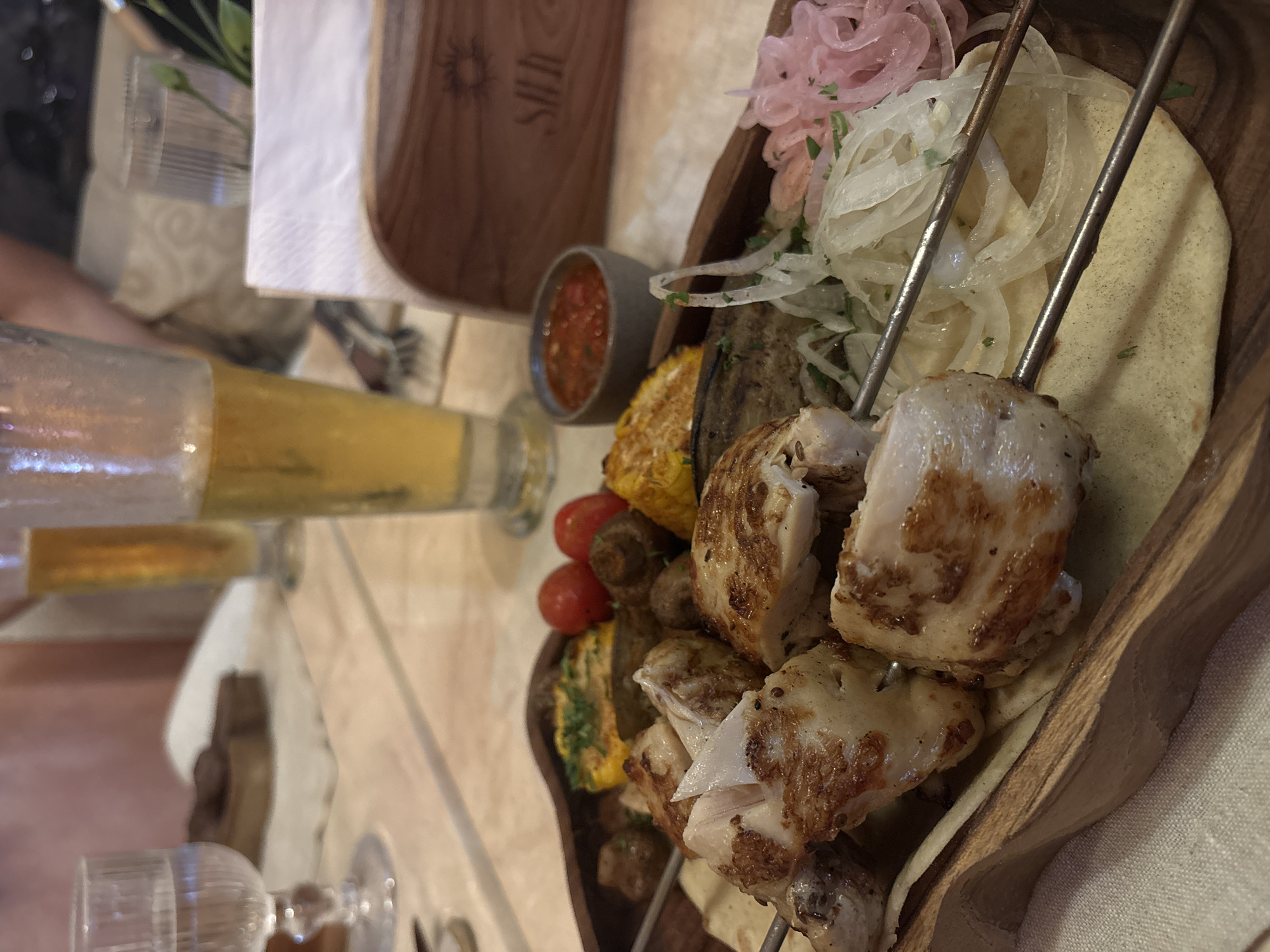 This image captures a close-up, slightly tilted view of a meal served on a wooden platter, likely at a restaurant in North Kuta, Indonesia. The main subjects are skewers of grilled meat, possibly chicken shashlik or kebab, cut into sizable chunks with a browned, crusty exterior and white, juicy interior. These are accompanied by grilled vegetables including bright yellow corn on the cob, green zucchini or eggplant slices, and vibrant red cherry tomatoes.
The skewers are laid over pieces of flatbread, possibly lavash or pita, which are topped with thinly sliced white onions and distinctively pink pickled red onions. A small grey ceramic bowl filled with a red, chunky sauce (perhaps a tomato salsa or adjika) sits beside the platter.
In the upper left, a partially visible hand holds a glass containing an amber-colored liquid, likely beer, suggesting someone is enjoying a drink with the meal. Another clear glass, possibly for water, is visible in the foreground, along with an empty wine glass further down.
The setting appears to be indoors, under warm artificial lighting, indicating an evening meal. A white paper napkin is folded neatly on the table. In the background, a tall, narrow wooden panel with a carved design and text is visible. The text appears to be "SIUI" or "SUIH" carved vertically, with a circular, stylized sun or flower logo above it. A small green plant in a clear glass adds a touch of decor. The overall atmosphere suggests a casual but pleasant dining experience.