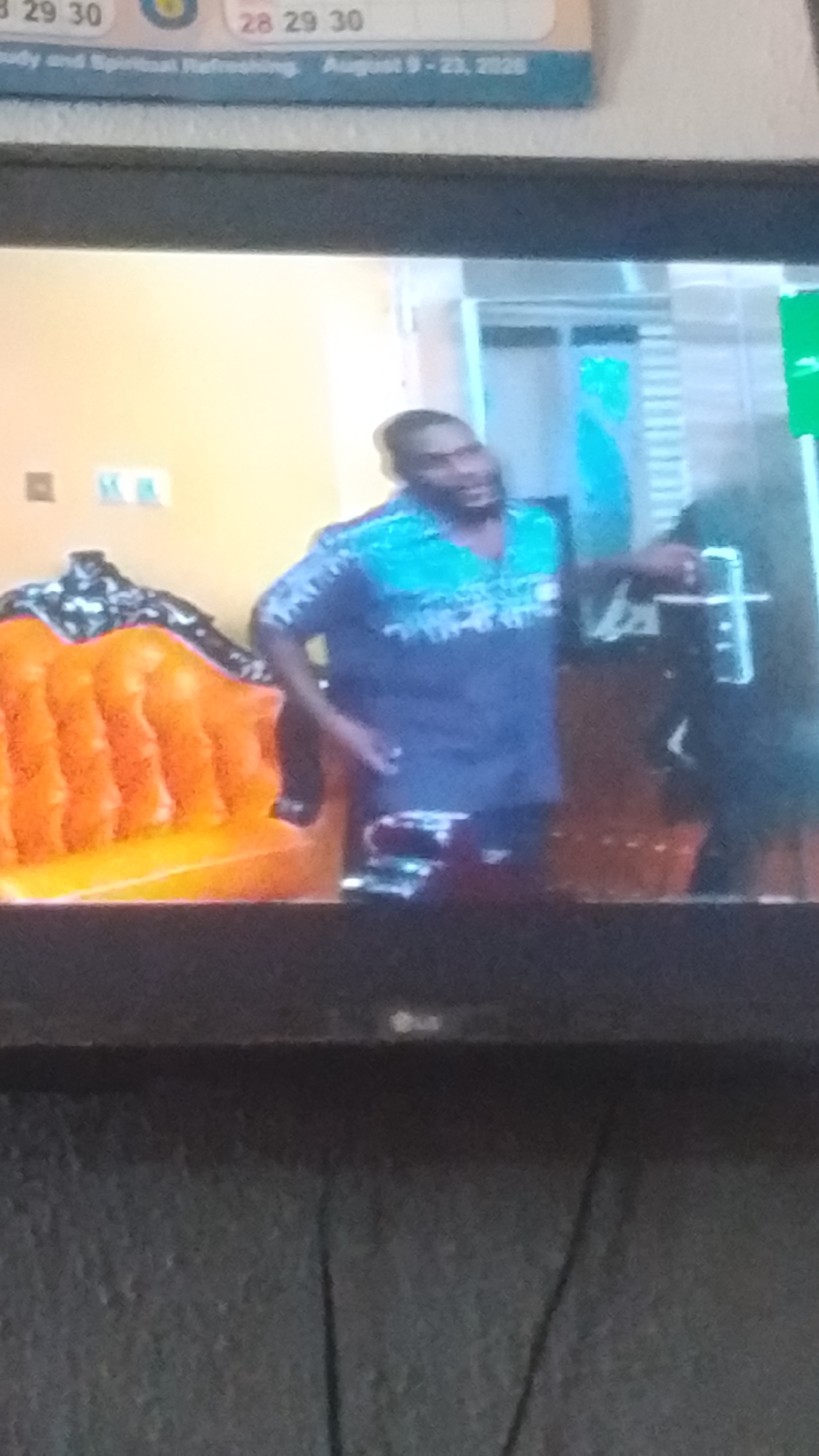 A man wearing a blue and teal patterned shirt stands to the right of a brightly colored orange sofa, partially visible on the left side of the frame. The man is looking to his left, with his right hand reaching towards a door frame, suggesting he may be entering or exiting. The background is blurry, but appears to be an indoor setting with warm-toned walls. The image is a photograph taken of a television screen, with the top edge of the television bezel visible at the top of the frame, and the textured surface of another screen or wall below.