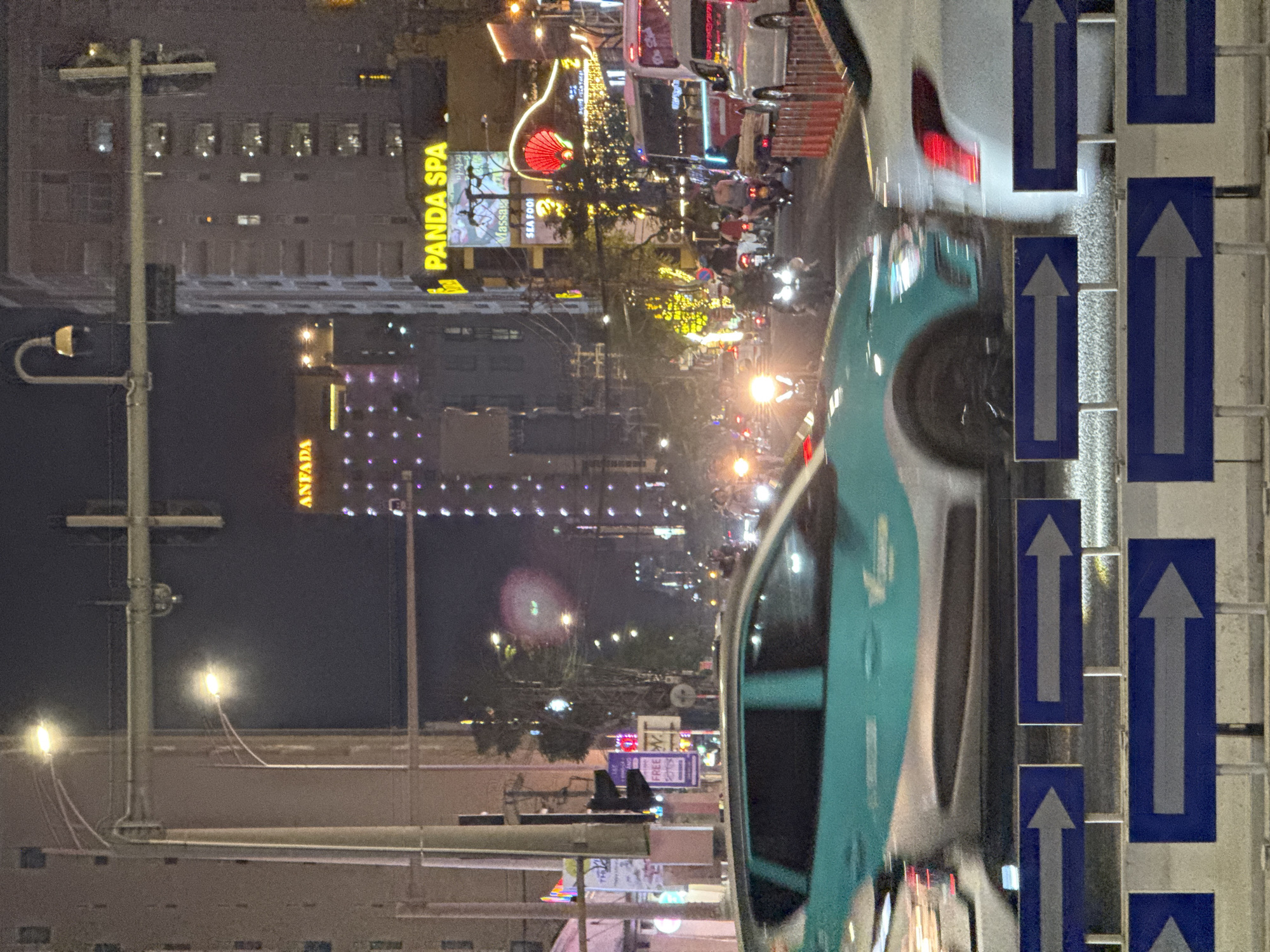 This nighttime image captures a vibrant urban street scene in Đà Nẵng, Vietnam, viewed from a slightly elevated perspective. The main subjects are a busy road, illuminated buildings, various vehicles, and street furniture.
In the foreground, the paved road shows signs of traffic, with a turquoise and white car, possibly a taxi, prominent on the right. Its design suggests a ride-hailing vehicle. Further ahead, other vehicles, including several motorcycles, can be seen, some with motion blur indicating movement. To the far right, a series of blue rectangular traffic signs with white upward arrows are vertically stacked, indicating a one-way street or lanes for forward movement.
The mid-ground is dominated by multi-story buildings adorned with numerous artificial lights, neon signs, and decorative illuminations. One large, bright yellow sign clearly reads "PANDA SPA." Below it, partially visible text suggests "Massag" (likely Massage) and "SEA FOO" (likely Sea Food). Another building further down the street features a yellow sign with "ANFAPA" (partially visible). Festive string lights and other glowing decorations add to the city's lively ambiance. Streetlights punctuate the scene, casting bright glows and streaks.
The overall impression is one of a bustling city at night, with a mix of commercial establishments and traffic. The dark sky contrasts with the extensive artificial lighting, creating a dynamic urban landscape common in Southeast Asian cities.
