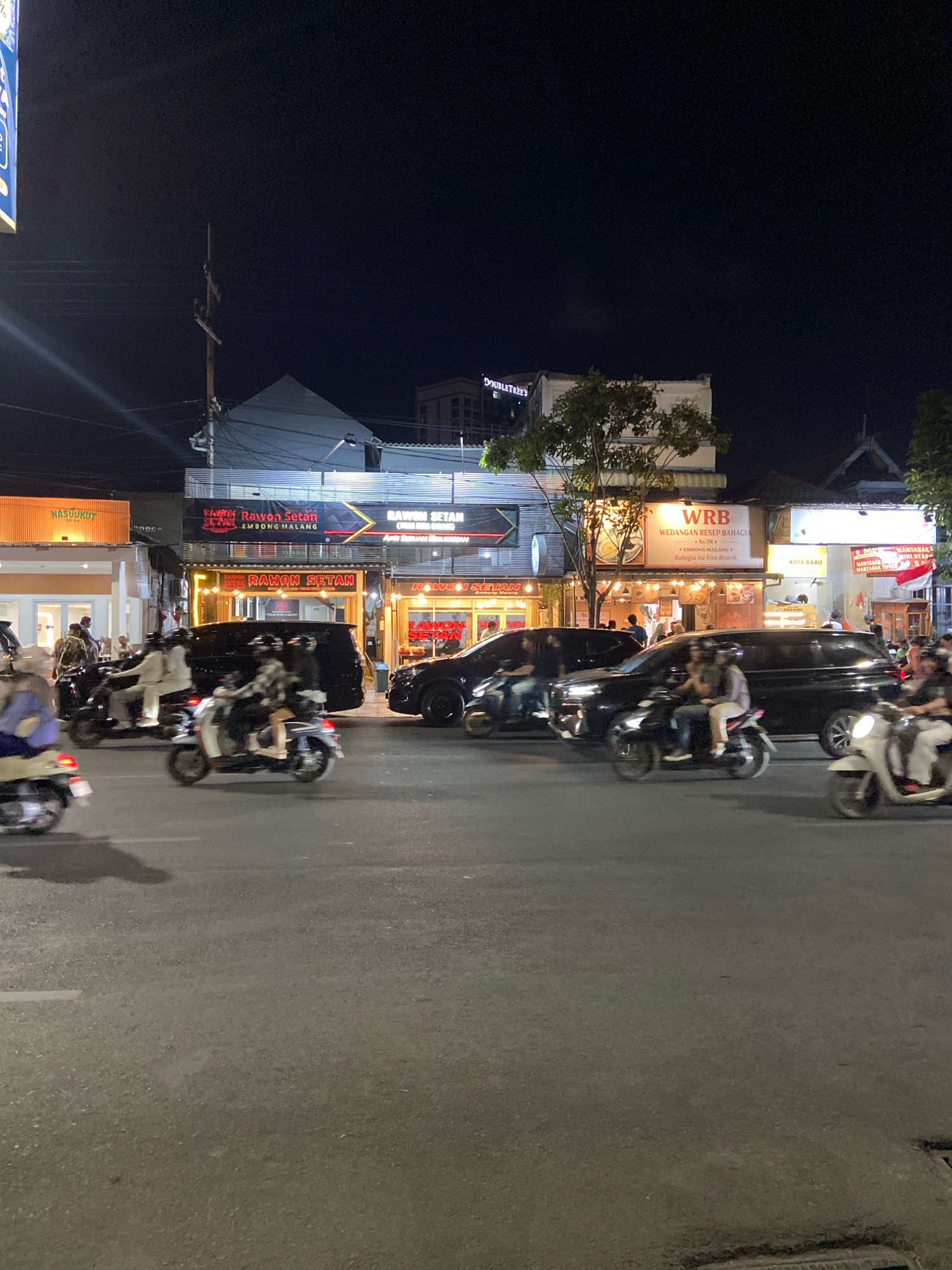 The image depicts a busy street scene at night in Surabaya, Indonesia, likely near the commercial district. The foreground is dominated by a road with several motorcycles and cars in motion, indicating traffic. In the background, a row of brightly lit shops and restaurants lines the street. Prominent signs include "NASUKUKET," "RAWON SETAN EMBONG MALANG," and "WRB WEDANGAN RESEP BAHAGIA," suggesting a vibrant culinary and commercial area. Buildings are illuminated, casting light onto the street and creating a lively atmosphere. A tall pole with electrical wires is visible, typical of urban infrastructure. The overall impression is one of a bustling nighttime urban environment.