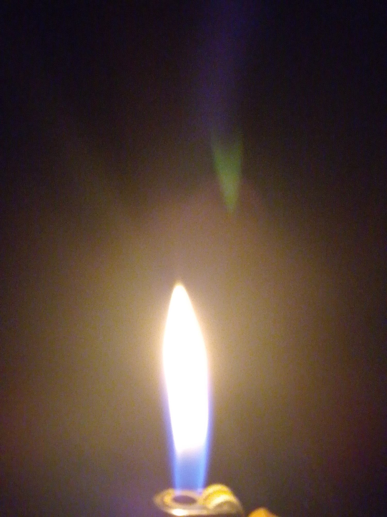 The image shows a close-up of a lighter flame in a dark environment. The flame is bright white at its base and transitions to blue, then to a pale yellow at the top, with a hazy glow surrounding it. The background is a dark, indistinct gradient of brown and black, with a faint greenish-purple light source visible in the upper background. The bottom edge of the image shows a portion of the lighter, including its metallic sparking mechanism. The scene does not provide any information that would allow for the determination of a location in Serti, Nigeria.