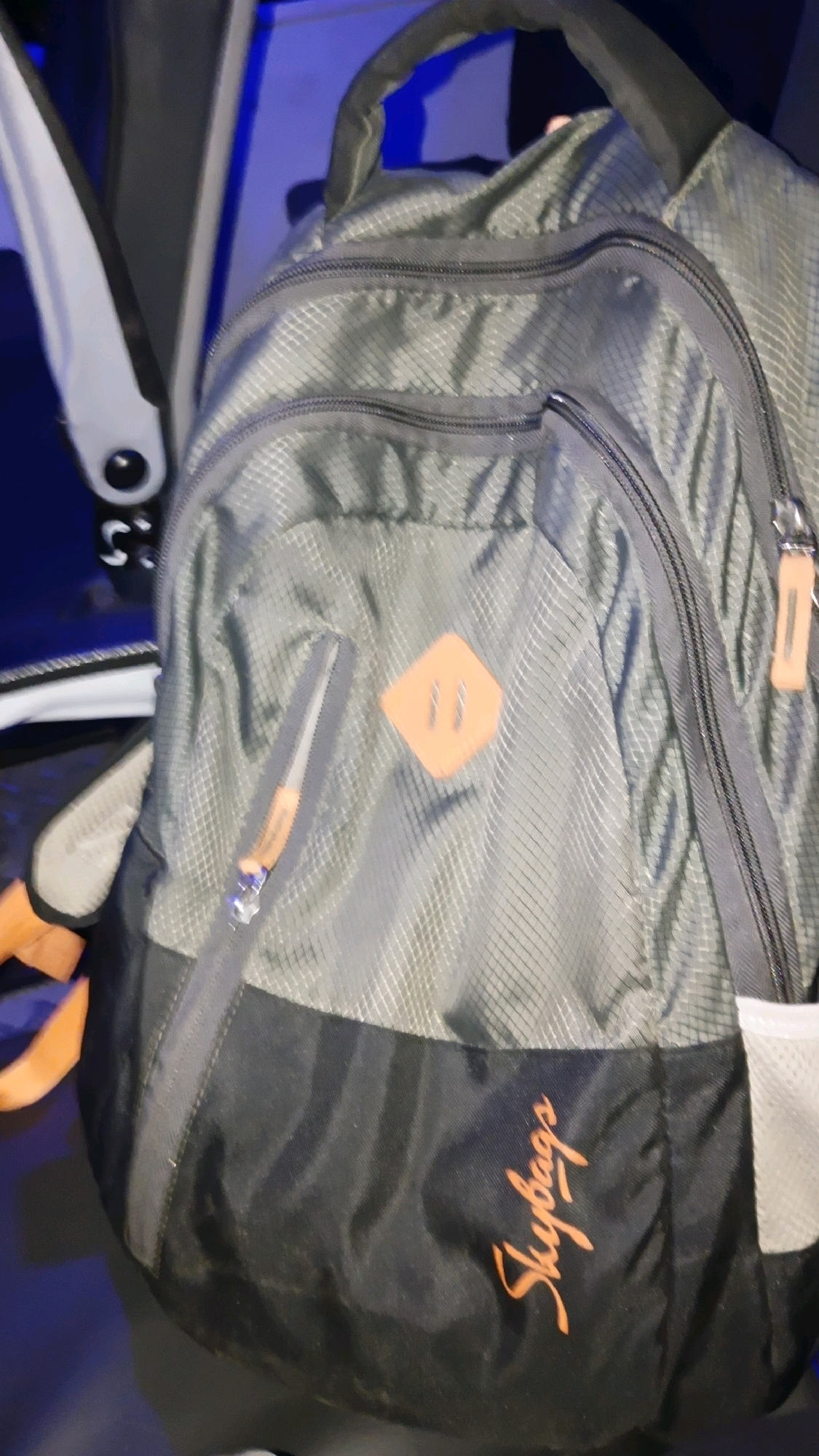 A grey and black backpack is displayed from a close-up, angled view. The backpack features multiple zippered compartments and a small orange square logo with two parallel lines on the front. The brand name "Skybags" is visible in orange script on the lower black section of the backpack. The background is blurred and shows hints of blue light and a white object to the left. The image appears to be taken indoors, likely in a domestic or storage setting. There are no people or animals visible in the frame. The scene is static and does not depict any action. The specific location in Karanji, India, cannot be determined from the visual information provided.