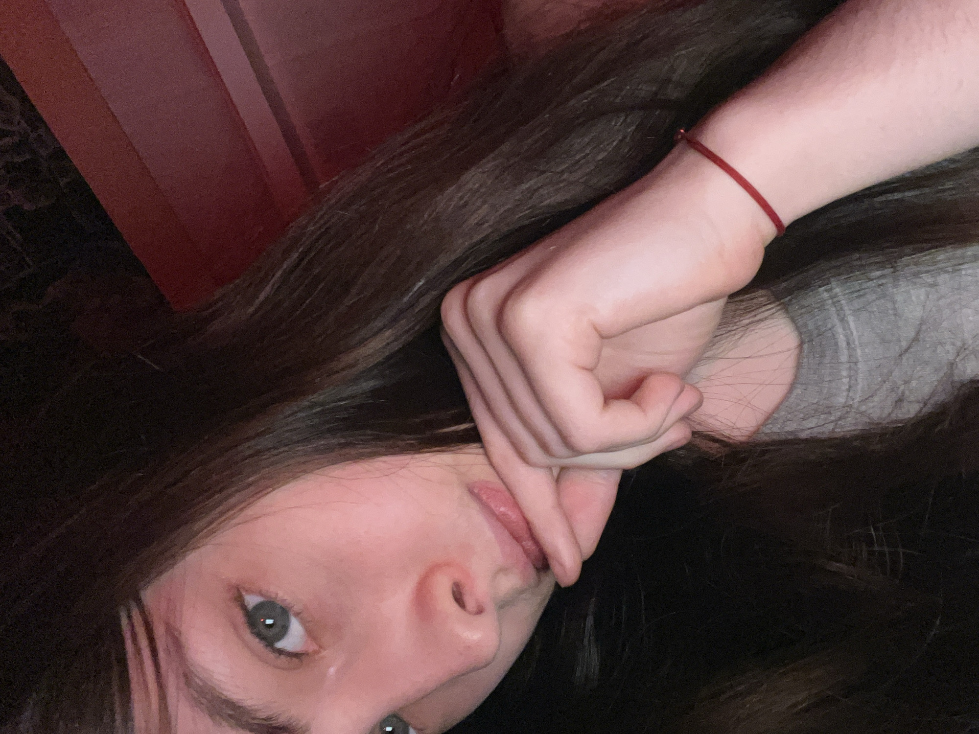 The image captures a close-up, diagonally oriented shot of a person, likely female, with long, dark brown hair. The subject is seen from a tilted perspective, possibly lying down, looking towards the camera. Her fair-skinned hand, adorned with a simple red string bracelet on the wrist, is positioned near her face, with an index finger gently resting on her lower lip, suggesting a pensive or shy demeanor.
Only a portion of her face is visible, including one grey-blue eye with a direct yet soft gaze, and her nose and mouth. Her lips appear full and naturally colored. A segment of a grey garment, possibly a t-shirt, is visible on her upper right arm and shoulder area.
The background is dimly lit, contributing to an intimate atmosphere. In the upper left corner, a dark red or maroon surface, possibly a wooden panel or piece of furniture, provides a structured element. The remainder of the background is dark, consistent with a low-light indoor setting. No specific location cues are visible beyond indicating an interior. There is no discernible text in the image.