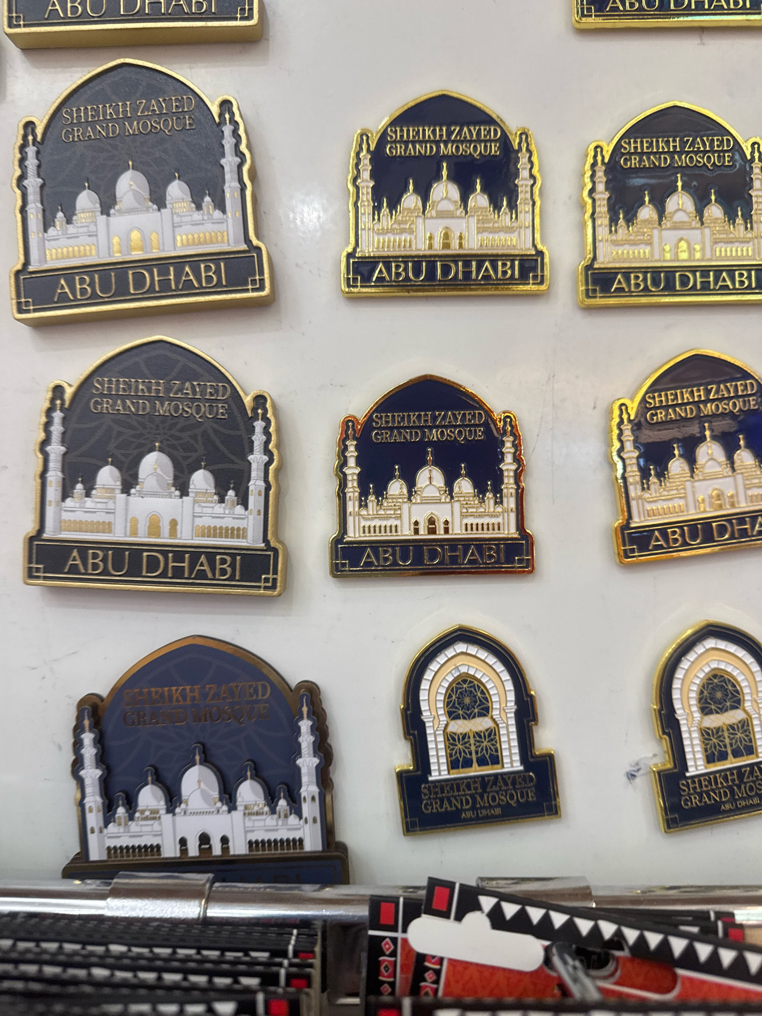 The image displays an assortment of refrigerator magnets, each featuring an illustration of the Sheikh Zayed Grand Mosque in Abu Dhabi, United Arab Emirates. The magnets are predominantly gold and dark blue or black, with depictions of the mosque's white domes and minarets. Some magnets also include the text "Sheikh Zayed Grand Mosque" and "Abu Dhabi". They are arranged in rows on a white surface, likely a wall or display board. In the foreground, the edge of what appears to be a display rack or shelf with black and red patterned objects is visible. The lighting suggests an indoor setting.
