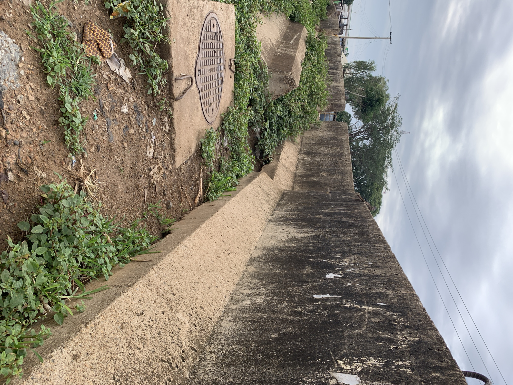 This image depicts an outdoor scene in Rafin Par, Nigeria, characterized by concrete infrastructure and dense vegetation under an overcast sky.
In the foreground and mid-ground, a textured concrete wall, approximately 1.5 to 2 meters in height, extends diagonally from the right into the distance. The upper portion of this wall displays significant dark discoloration, likely from moisture or organic growth, with scattered white remnants adhering to its surface. The lower section of the wall is a lighter, sandy-colored concrete, forming a distinct base. Parallel to this wall, a concrete drainage channel is partially visible at its base, largely obscured by thick green plant growth.
To the left of the main wall, a series of pre-fabricated or cast-in-place concrete structures, forming what appear to be open drainage channels or culverts, are visible. These structures are extensively overgrown with verdant vegetation, filling much of their internal space. In the immediate foreground, slightly left of center, a circular, dark-colored cast-iron manhole cover with a grid pattern is set into a square concrete slab. Two U-shaped metal bars are embedded in the concrete slab near the manhole cover.
The ground consists of exposed, reddish-brown dirt, interspersed with numerous patches of low-lying green weeds and grasses. Scattered debris, including a piece of patterned brown fabric and various small fragments of plastic or paper, is visible on the ground surface.
In the background, above the concrete structures, several large green trees are prominent. Utility poles with overhead power lines are visible in the distant left, and a faint elevated structure, possibly a bridge or road, can be discerned further back. A slender tower or antenna is also faintly visible against the cloudy sky in the distant background. The sky is entirely covered by a thick layer of grey clouds, indicating no direct sunlight. No individuals or active interactions are present in the scene.
