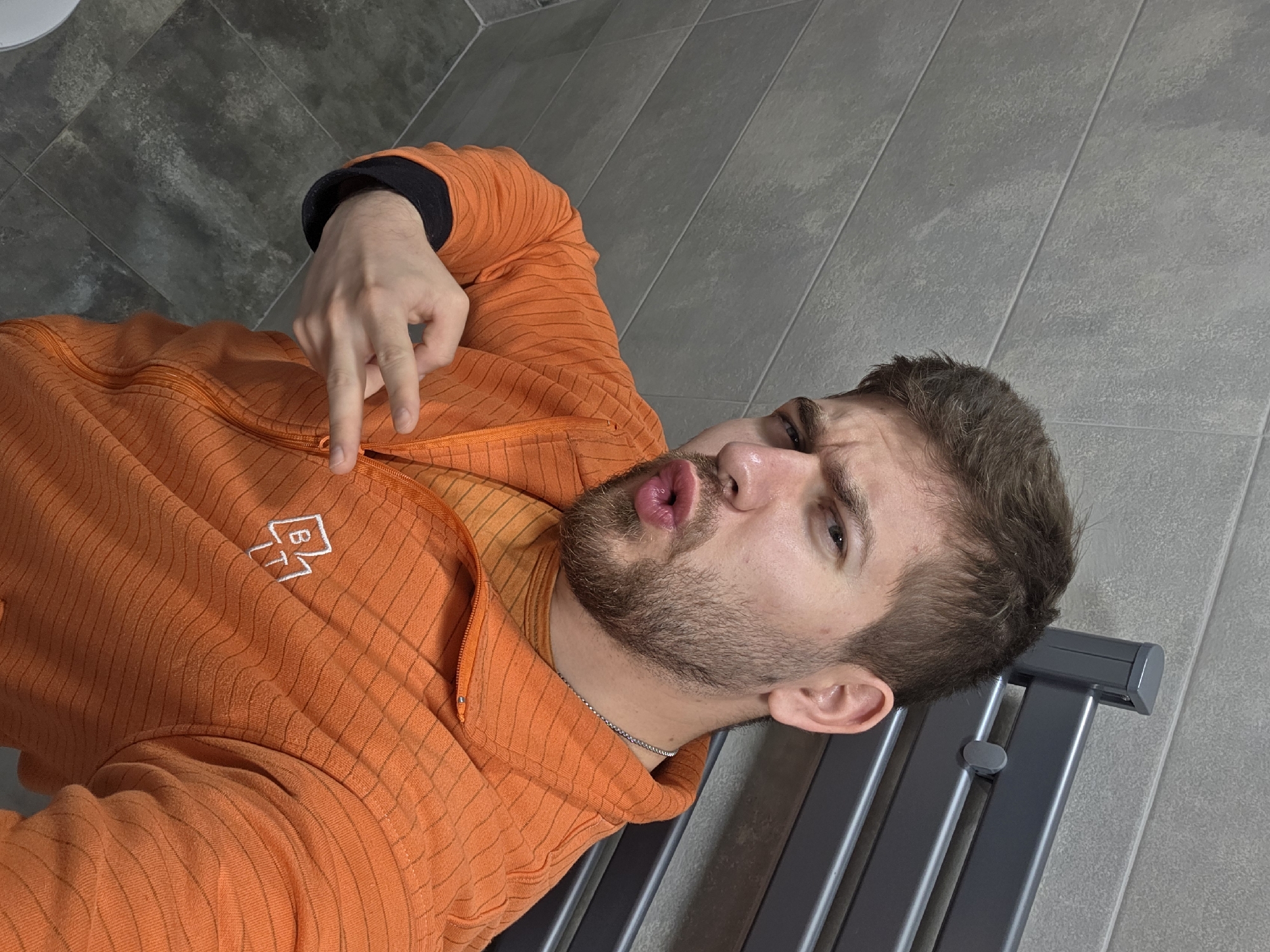 A medium shot depicts a man lying on a gray towel rack. He is wearing an orange zip-up hoodie with a white logo in the top left corner. His left hand is raised, making a peace sign. He is pouting his lips. The background is gray tiled walls. The location is unknown and not specified as Yagodovo, Bulgaria.