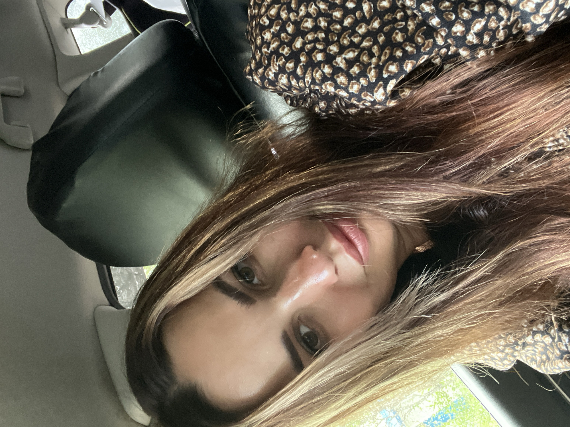 A medium shot shows a woman in the passenger seat of a vehicle. The woman has long, wavy brown hair with blonde highlights, and she is wearing a leopard print top. She is looking at the camera with her head tilted to the side. The background is blurred, but it appears to be a rainy day outside the car window, with trees and buildings visible. The interior of the car is dark and appears to be a typical passenger car.