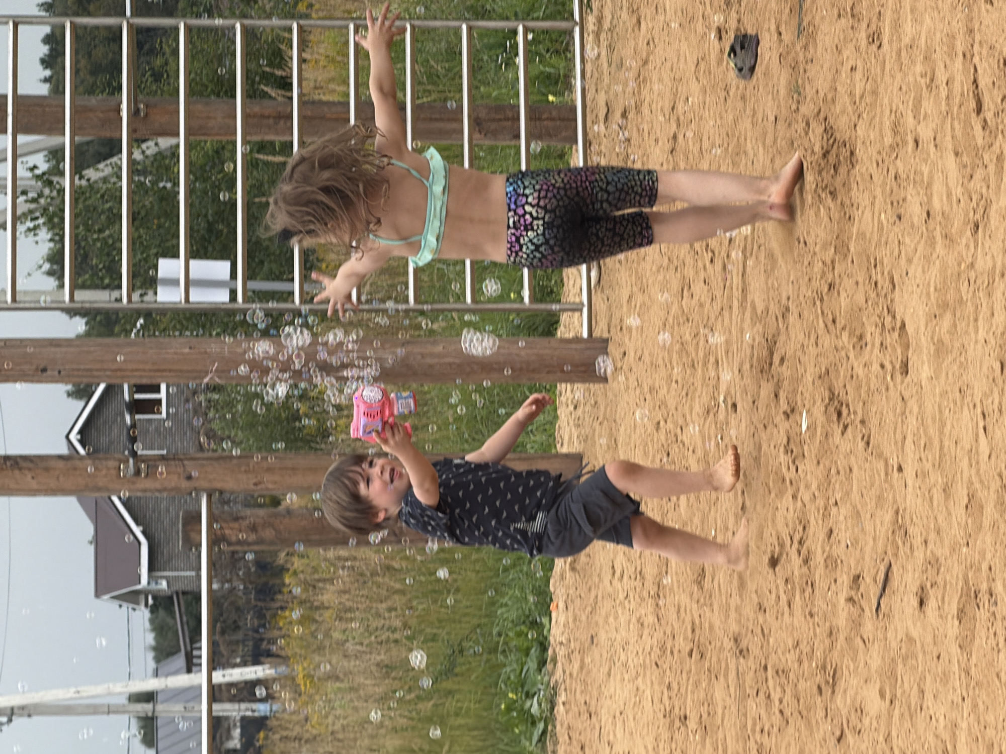 In a sandy outdoor setting, two children are actively playing. On the right, a young girl is in mid-air, her body arched backward as if jumping or falling. She is wearing a light blue bikini top and patterned shorts with a leopard-like print. Her arms are outstretched, and her hair is flowing. On the left, a younger boy is looking up and holding a pink bubble gun, from which numerous bubbles are emanating. He is wearing a dark t-shirt with a small triangle pattern and dark shorts. The background features a wooden structure with vertical slats, some greenery, and buildings, suggesting a residential area. The ground is covered in sand, with some grass visible in the background. The scene appears to be during the daytime, likely with clear or partly cloudy weather, given the lighting. The children's expressions and energetic movements suggest joyful play and a sense of fun. The presence of a bubble gun and the falling bubbles indicate a playful activity.