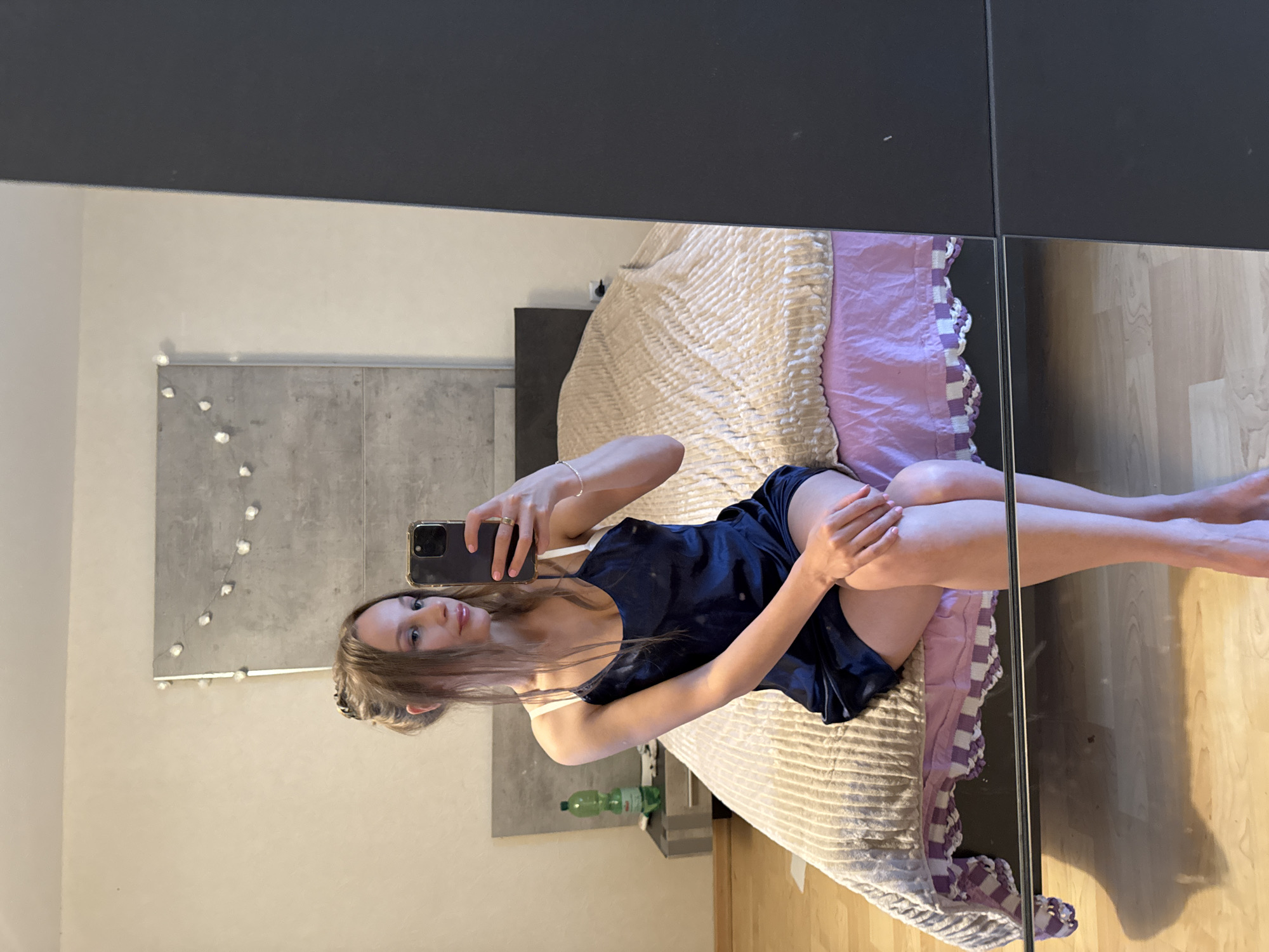A young woman is sitting on a bed in a bedroom. She is wearing a dark blue slip. She is holding a smartphone in her right hand and taking a mirror selfie. The bed has a beige comforter with a textured pattern and a pink bed skirt. To the left of the bed is a nightstand with a water bottle on it. The room has light-colored walls and a wooden floor. The image is a photograph taken in Überherrn, Germany.