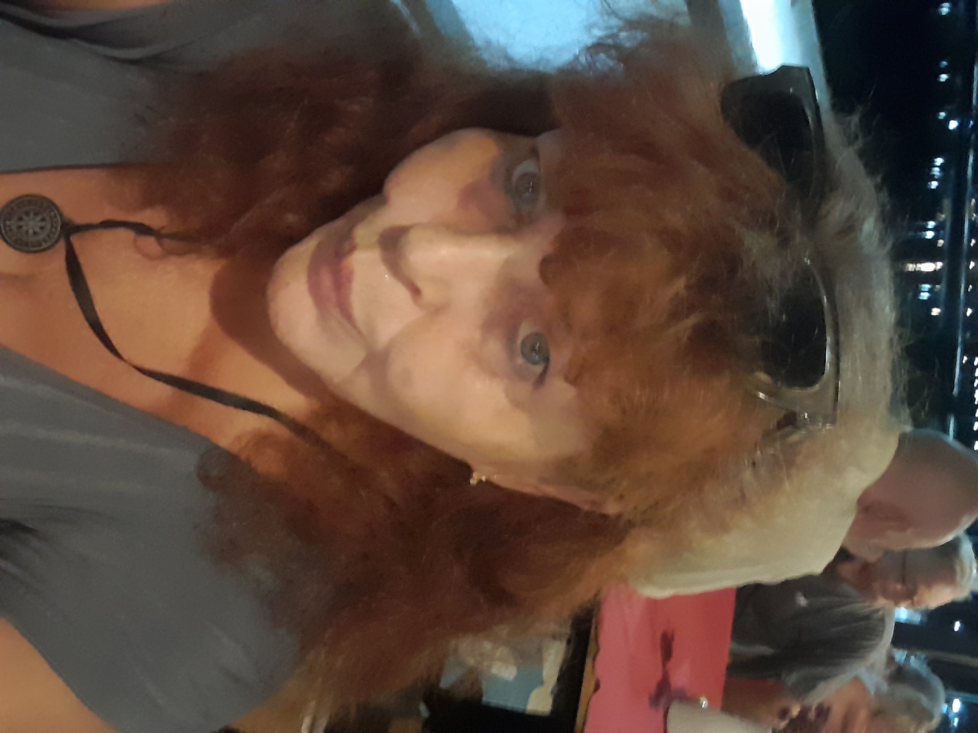 A woman with reddish-brown hair and sunglasses on her head faces the camera. She wears a grey top and a necklace with a round pendant. Behind her, two elderly people are visible in the background; one has glasses and is wearing a grey shirt. The background has bokeh lights, suggesting a night scene. It is uncertain that this picture takes place in the city of บ้านบางรักษ์, Thailand.