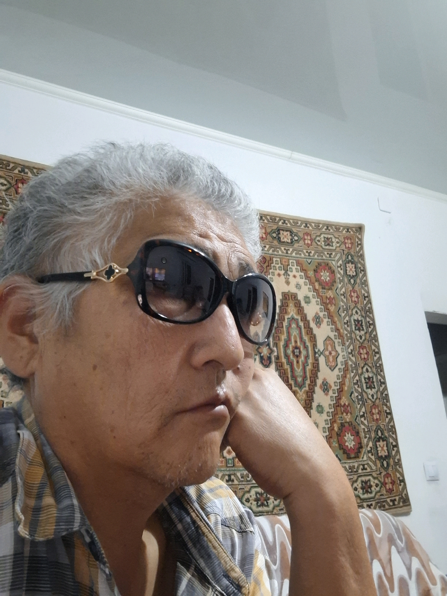 A close-up, medium shot captures a person with graying hair and sunglasses indoors. The individual, positioned in the center of the frame, is looking off to the right with their hand resting on their chin. They are wearing a plaid shirt. Behind them, a patterned rug hangs on a light-colored wall. The overall setting suggests a domestic environment, possibly a living room. The image does not contain sufficient detail to determine the exact location within Almaty, Kazakhstan.