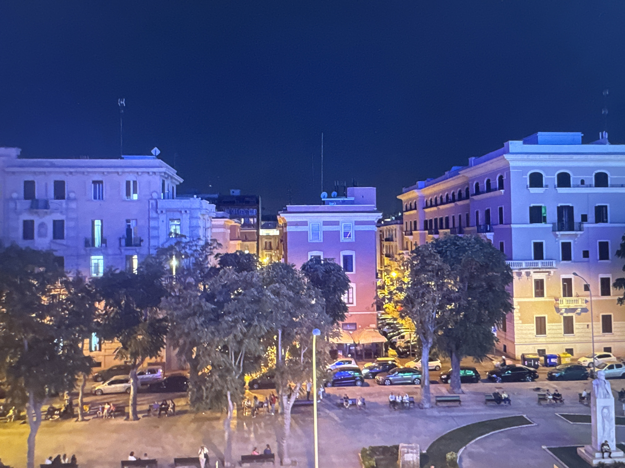 The image captures an urban scene at night, likely from an elevated perspective. The sky is a deep, dark blue, indicating nighttime. In the foreground, a paved area with curved green borders is visible, with several people seated on benches. A tall, slender monument or sculpture stands to the right, with a single person seated at its base. Beyond this, trees with dark foliage line the area, with some illuminated by streetlights.
In the mid-ground, a street is lined with vehicles, including a white sedan, a blue hatchback, and several dark-colored cars, one of which is a dark green SUV. Buildings with illuminated windows and architectural details are prominent. On the left, a light pink building features multiple floors with windows and balconies. To its right, a bright pink building has a prominent window on its upper level and lights on its lower floors, suggesting commercial establishments. Further to the right, a cream-colored building exhibits a row of arched windows on its upper floors and balconies. Several streetlights illuminate the area.
The background consists of more buildings, some with illuminated windows, and the dark night sky. The overall impression is of a city square or street scene at night, with a mix of residential and commercial buildings, public spaces, and vehicular traffic. The image does not contain any elements that suggest the location is Porrentruy, Switzerland.