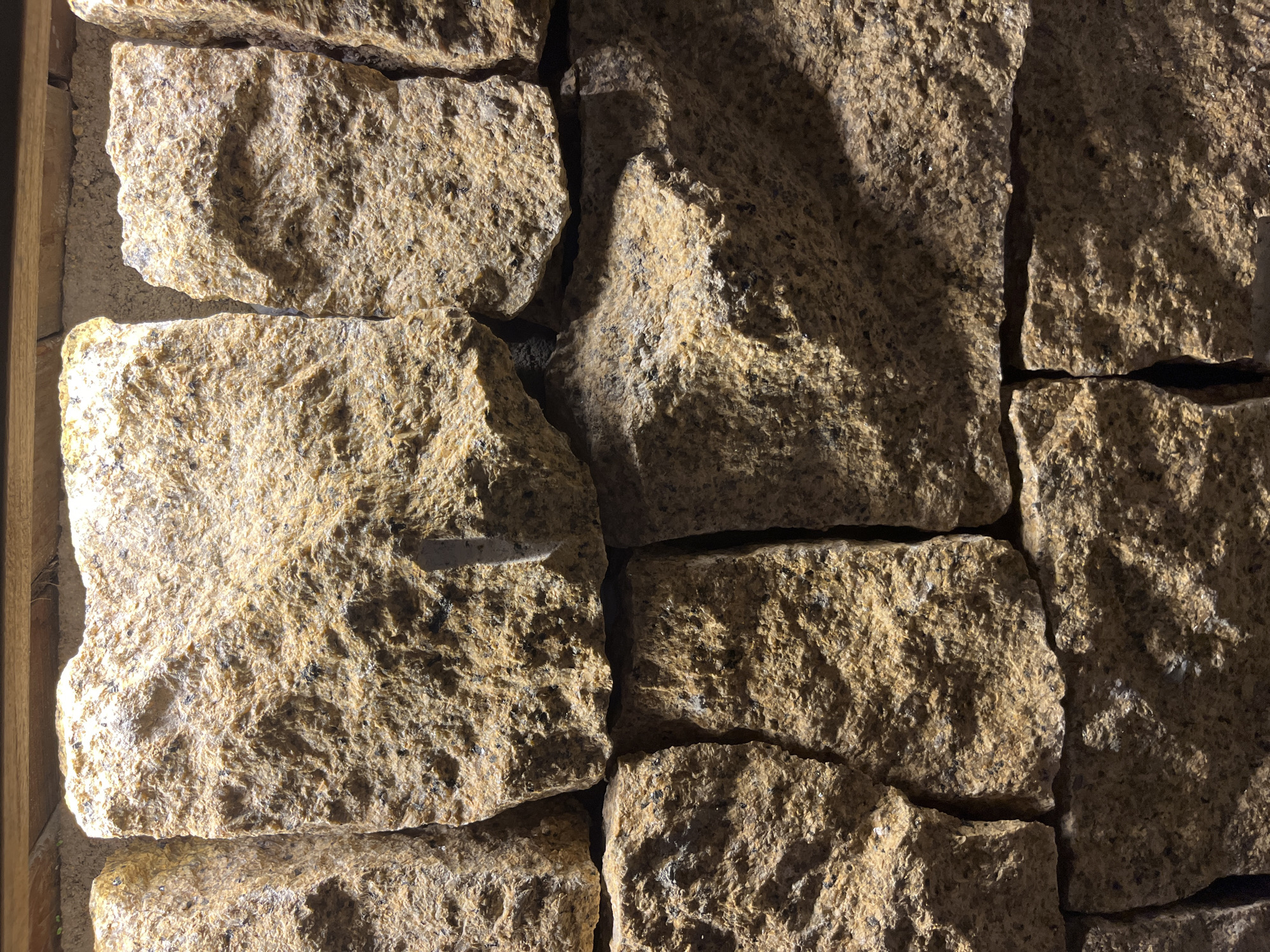 The image is a close-up, vertically oriented shot of a rustic stone wall. The stones are rough-hewn, irregular in shape, and vary in size, with earthy tones of brown, tan, and flecks of dark gray and black. They are stacked tightly together, creating a textured surface. The lighting appears to be artificial and directional, casting strong shadows and highlighting the rough texture of the rocks. The top of the image shows a wooden beam or frame. There are no people or animals present, and no specific actions are depicted. The background is entirely the stone wall itself. The scene is likely an outdoor or interior decorative wall. The prompt asks for the location to be "Bac Nha Trang Ward, Vietnam", but there is no visual information in the image to confirm or deny this specific location.