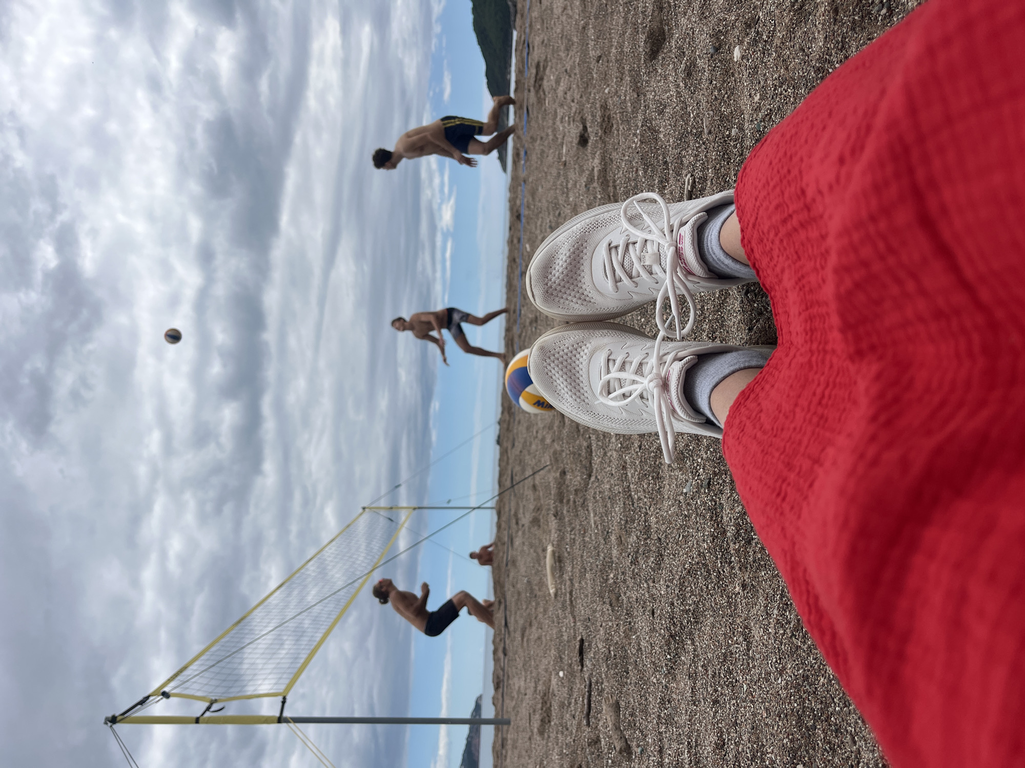 From a first-person perspective on Boreti beach, Montenegro, the image captures a beach volleyball game in progress. Three shirtless men in dark shorts play near a yellow volleyball net. A volleyball is in mid-air. White sneakers and a red garment are visible in the foreground, resting on the dark sand. The sky is overcast. A mountain range can be seen in the background.