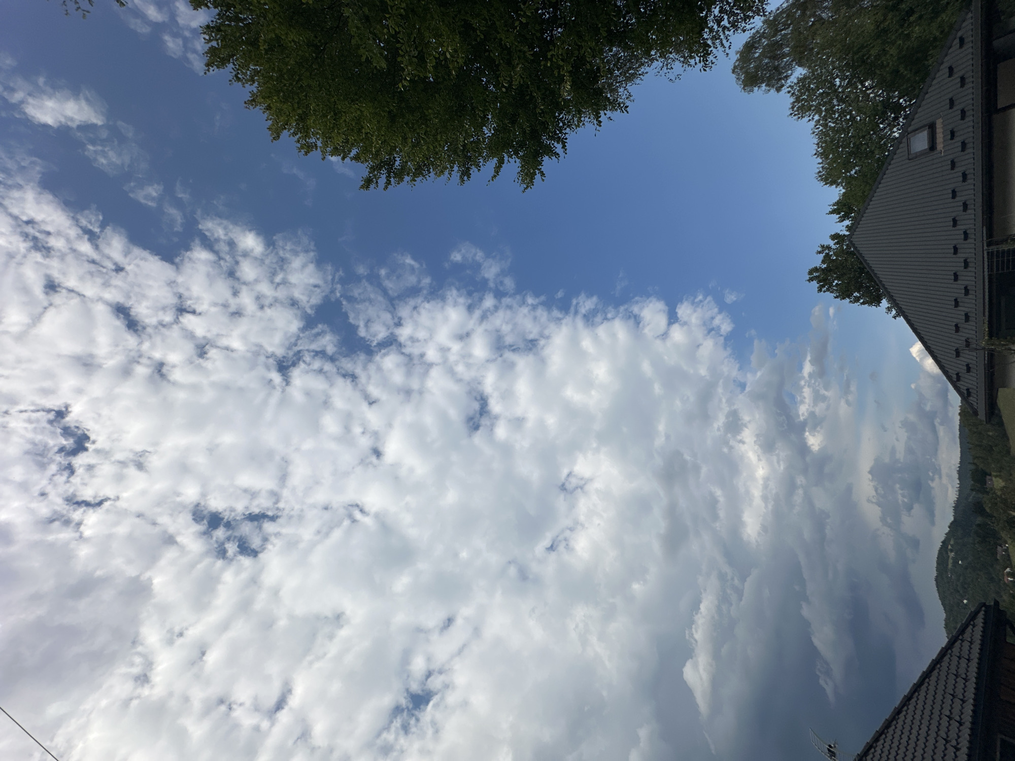 The image captures a view from a residential area in Wisła, Poland, looking upwards towards the sky. A significant portion of the sky is filled with a dramatic display of white and dark grey clouds, suggesting a change in weather, possibly the approach of a storm. The clouds are dense and textured, with patches of bright blue sky visible on the right side of the frame.
In the foreground, the dark, sloped roofs of two houses are visible. The house on the left has a dark, textured roofing material and appears to be partially in shadow. A portion of what might be a satellite dish or antenna is visible on the roof's edge. The house on the right features a dark, corrugated metal roof with a small, square skylight.
Beyond the houses, a dense green forest covers rolling hills. The trees are lush and varied in shape, indicating a natural, possibly mountainous landscape characteristic of Wisła. The overall impression is one of a rural or semi-rural setting with a dynamic sky overhead. There are no people or animals visible in the image.