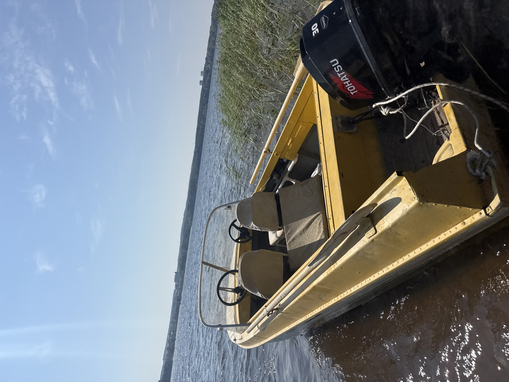 A yellow motorboat is docked near reeds on a sunny day. The boat has two seats covered with beige fabric, a steering wheel, and a black Tohatsu outboard motor with a "30" visible. In the background, across the water, a treeline with some distant buildings is visible under a clear blue sky with scattered clouds. The water is dark and slightly rippled. The scene appears to be on the Kama River near Chaikovsky, Russia.