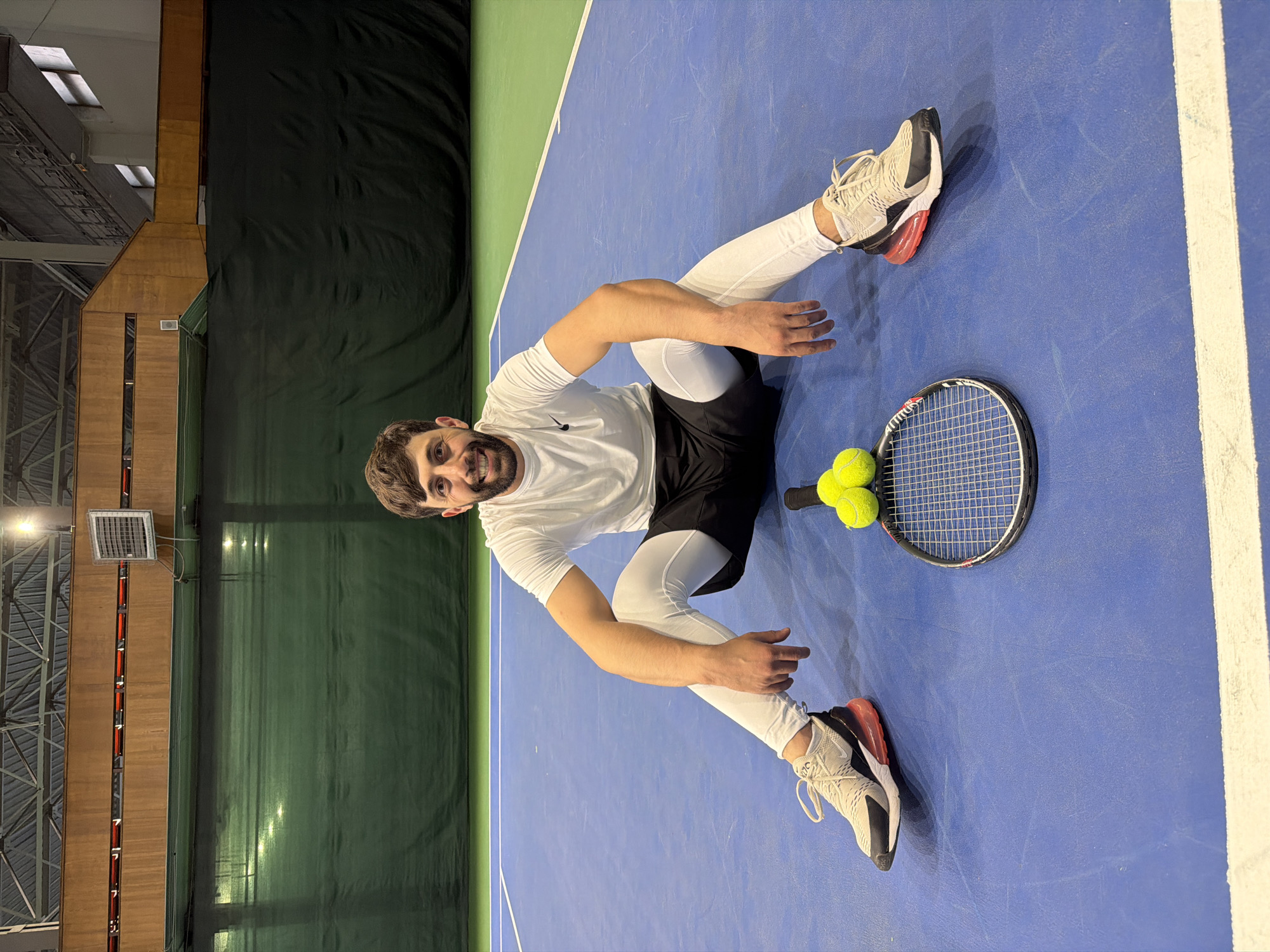 A man is sitting on an indoor blue tennis court, legs outstretched in a wide V-shape. He is wearing a white t-shirt, black shorts, white leggings, and beige and black sneakers with red accents. He is smiling at the camera. Next to him is a tennis racket with two tennis balls resting on the strings. The court has white lines and a green wall. The location could be the Yenisei Tennis Hall in Krasnoyarsk, Russia. The indoor sports complex has a high ceiling with metal beams, industrial lighting fixtures and curtains.