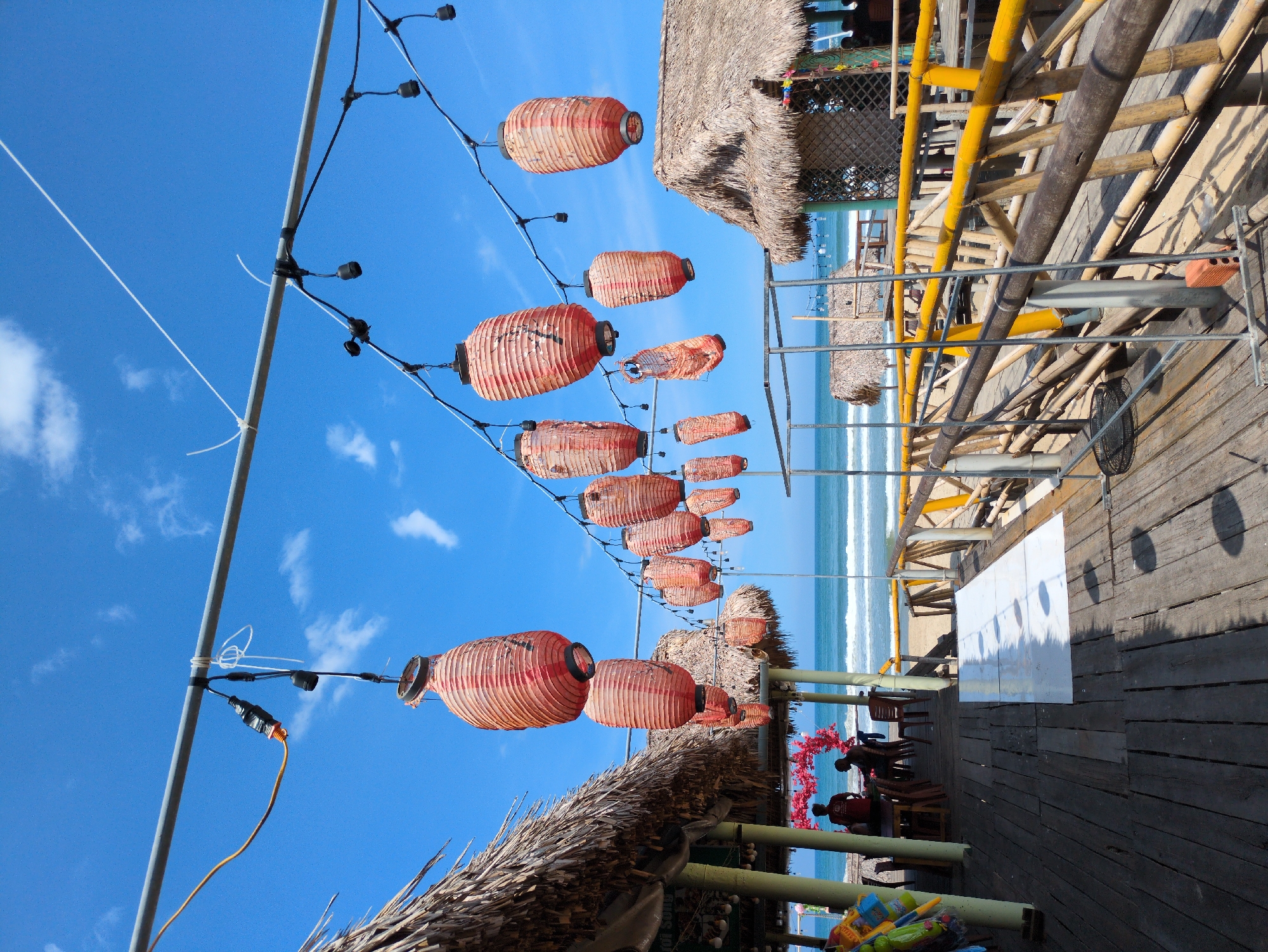 This image depicts a beachfront scene in Huế, Vietnam, featuring a wooden deck with thatched-roof structures and a string of red lanterns hanging overhead. The deck is furnished with tables and chairs, suggesting a dining or relaxation area. Beyond the deck, the ocean stretches out under a bright blue sky dotted with scattered clouds. The overall atmosphere appears to be that of a seaside establishment, likely a restaurant or cafe, capitalizing on its scenic coastal location. The lanterns, a common decorative element in Vietnamese culture, add a vibrant and traditional touch to the otherwise natural setting.