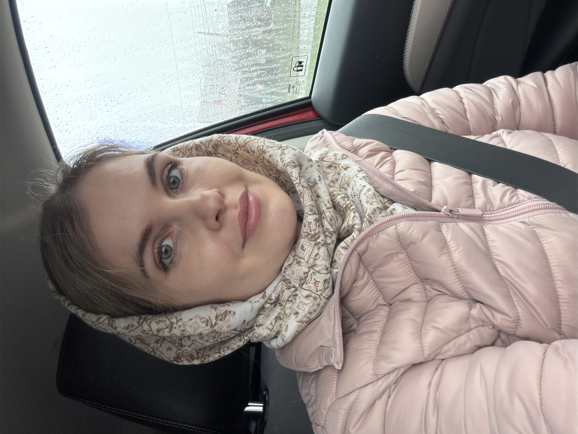 A woman with fair skin and green eyes is sitting in a car, wearing a light pink puffer jacket and a patterned scarf or head covering. She has a seatbelt fastened across her chest. Outside the car window, it appears to be raining, with water droplets visible on the glass. The interior of the car includes a dark seat and a red door frame. The location is not discernible as Izhevsk, Russia based on the provided image.