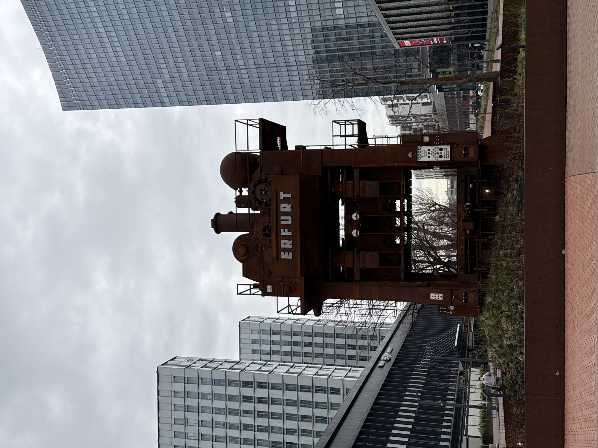 The image shows a large, rusted metal industrial press with the word "ERFURT" prominently displayed on its side. It's situated outdoors, likely as a monument or art installation. The press is surrounded by modern buildings, including a large glass-paneled skyscraper. A black staircase is visible, and some vegetation is present in the ground around the press. There is one person wearing a white jacket walking near the stairs. The location is the White Square Business Center in Moscow, Russia. The sky is overcast.