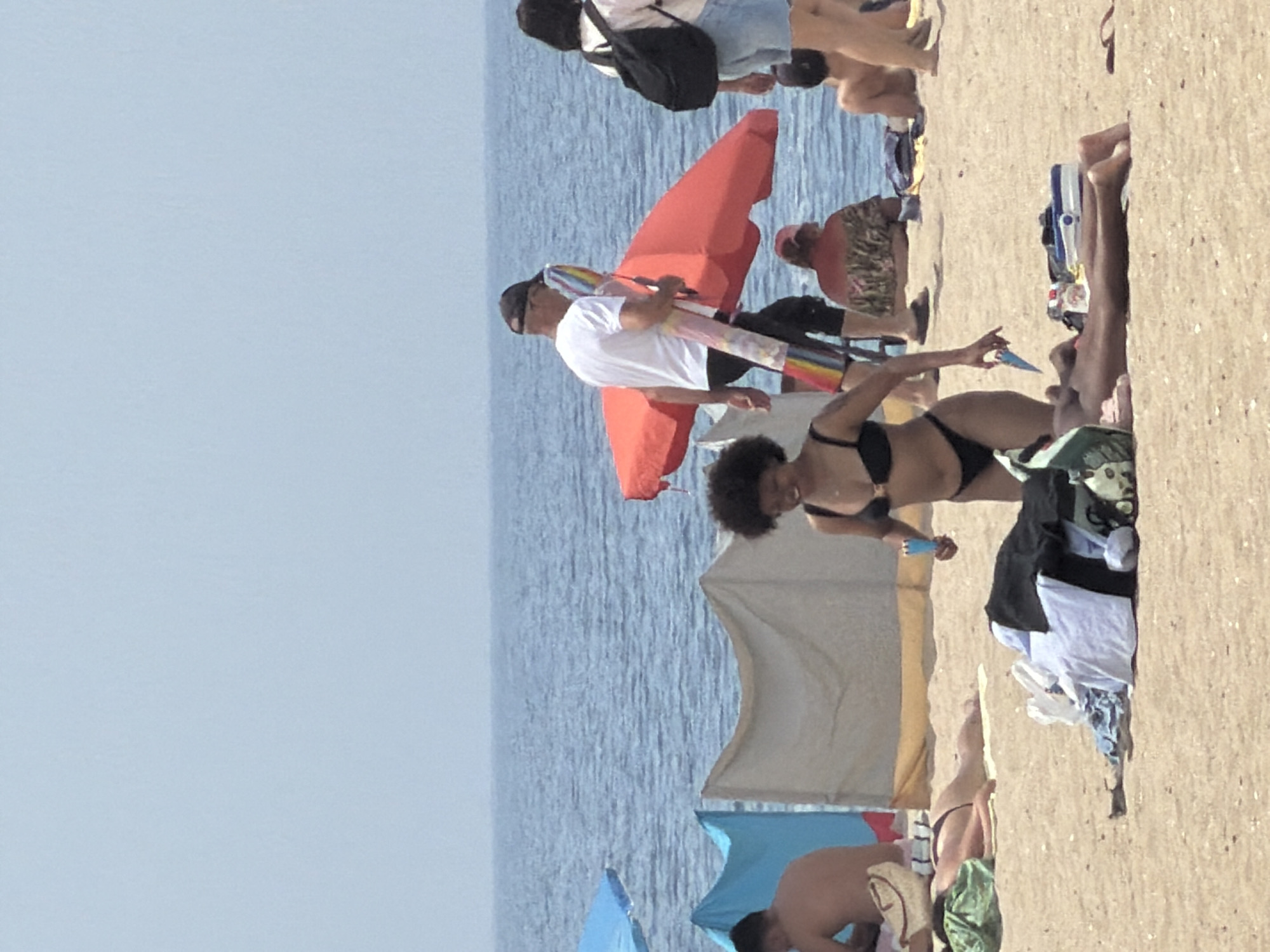 The image depicts a sunny day at a beach, likely in Espinho, Portugal, given the context of a previous request. In the foreground, a woman with dark, curly hair is wearing a black bikini. She is holding a blue object, possibly an ice pop or a toy, in her right hand and reaching towards the sand with her left. A pile of clothes lies on the sand in front of her. Behind her, another person is lying on the sand with a green towel draped over their legs. Further back, a man in a white t-shirt and black shorts, wearing a dark baseball cap, is walking and carrying a rolled-up colorful item under his arm. He is near a large red umbrella. To the left, a blue beach umbrella is partially visible. In the background, the blue ocean stretches to the horizon under a pale blue sky, with several other beachgoers and beach shelters scattered across the sandy shore.