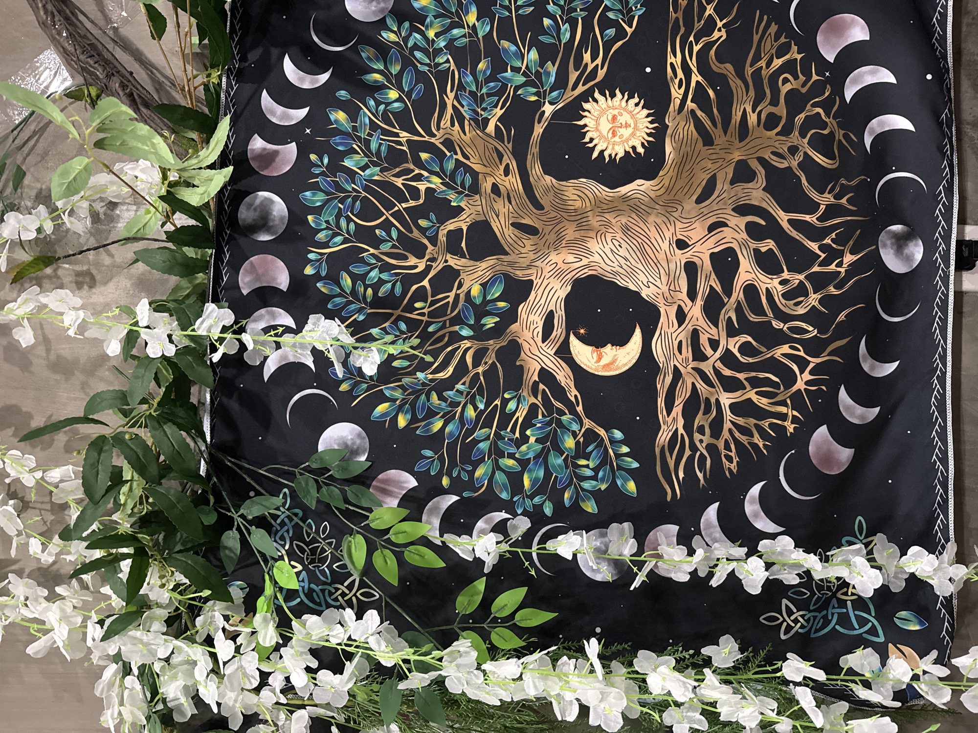 The image depicts a close-up, overhead view of a black tapestry featuring a gold and green tree of life. The tree's roots are prominent, spreading across the bottom of the tapestry. Various phases of the moon, depicted as circles and crescents in shades of gray and pink, are arranged in a repeating pattern along the left and bottom edges. To the upper left of the tree, a crescent moon with a face is visible, and to the upper right, a radiant sun with a face is depicted. The tapestry is partially obscured by cascading artificial white flowers and green leaves, which appear to be part of a decorative arrangement. The background is a plain, textured wall. There are no people or animals present in the image. The scene is indoors and does not provide any specific clues about its location within Đà Nẵng, Vietnam.