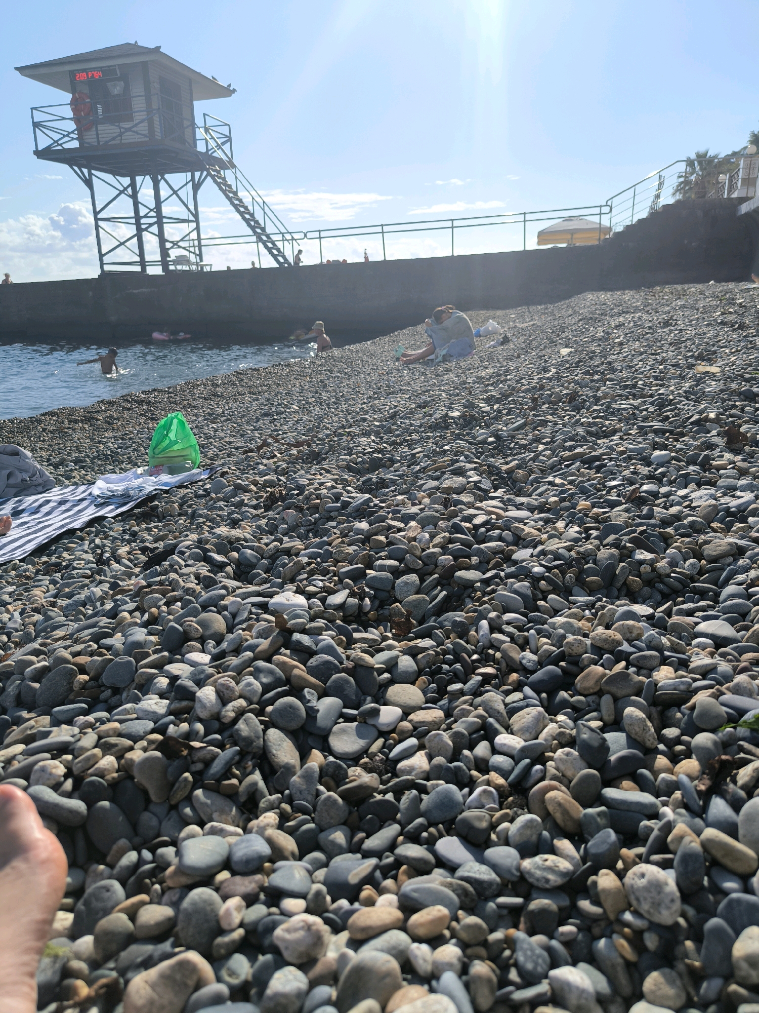The image depicts a pebble beach in Sochi, Russia, on a sunny day. In the foreground, a person's foot is visible on the stones. Further back, a striped towel and a green plastic bag are laid out on the pebbles. Several people are visible in the shallow water, with some playing and others swimming. In the background, a lifeguard tower stands prominently on a concrete structure overlooking the sea. The sky is bright blue with some scattered clouds, and the sun is shining, casting a glare on the scene. The overall atmosphere suggests a leisurely day at the beach.