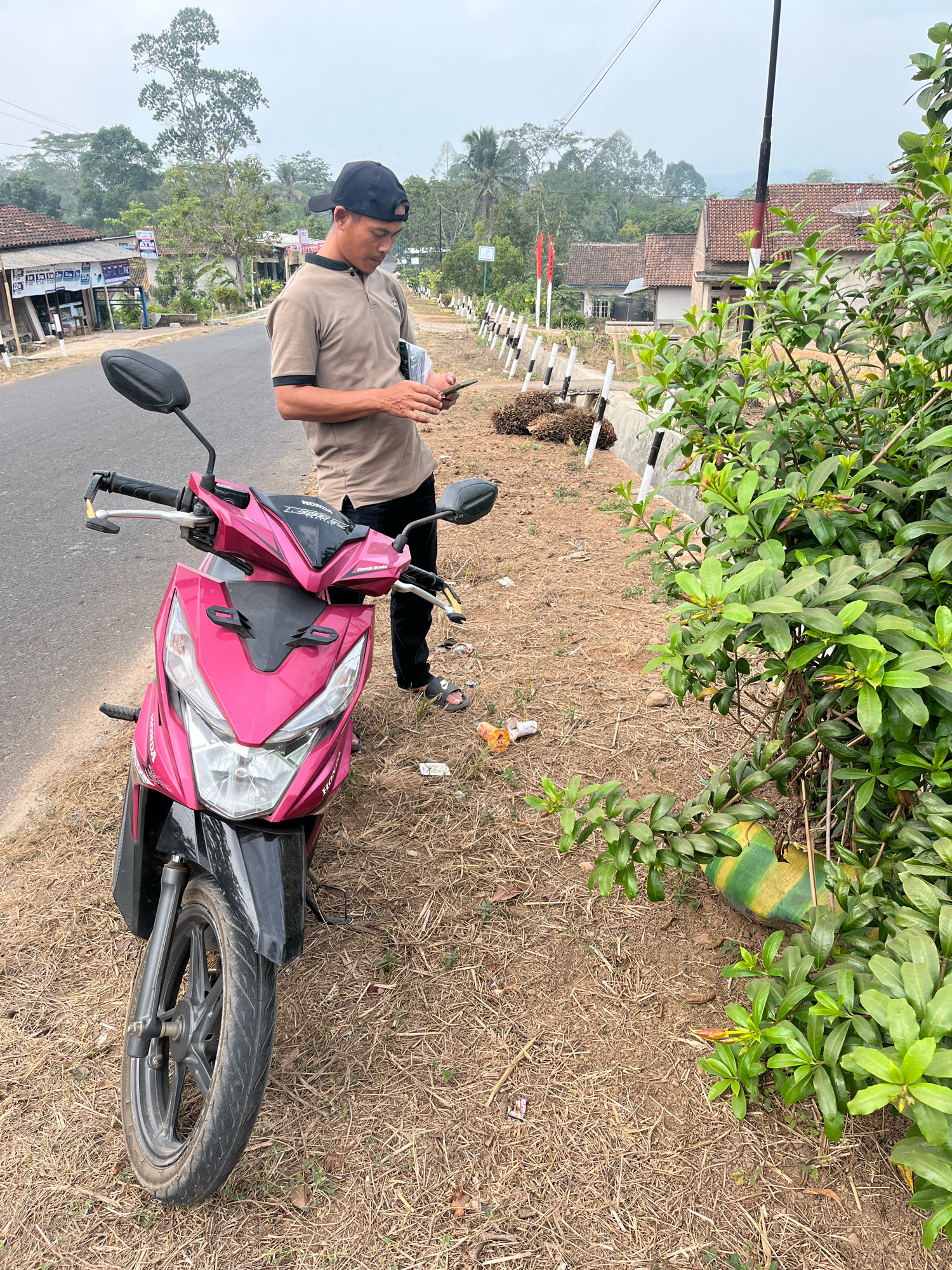A man wearing a brown polo shirt, black cap, and dark pants stands by a pink and black motorcycle parked on the side of a paved road in Muara Dua, Indonesia. The man is looking down at his phone. The motorcycle is facing left, and its front wheel is close to the edge of the road. The area next to the road is covered in dry grass and dirt, with some scattered debris. In the background, there are houses with tiled roofs, trees, and a cloudy sky. A line of white posts with black stripes runs along the edge of the road, separating it from a grassy embankment. Some palm fronds are piled near the posts.