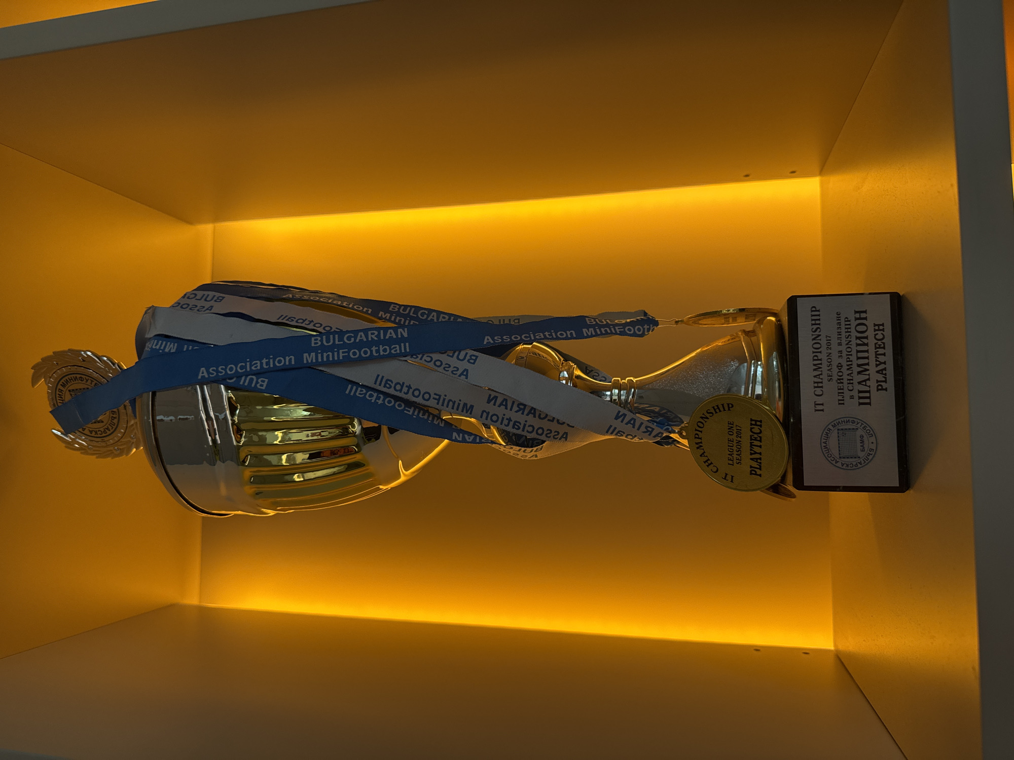 An eye-level shot captures a silver-toned trophy and associated medals displayed on a shelf, illuminated by warm yellow LED strip lighting from below. The trophy is oriented horizontally, adorned with multiple blue ribbons or straps that bear text.
Visible text on the blue ribbons, some mirrored, includes "BULGARIAN Association MiniFootball" and "Association MiniFootball". A circular medal-like attachment on the trophy reads "IT CHAMPIONSHIP," "LEAGUE ONE," "SEASON 2017," and "PLAYTECH."
To the right of the trophy, a rectangular award plaque stands vertically. Its text is primarily in Bulgarian and English. The English text states "IT CHAMPIONSHIP" and "SEASON 2017." The Bulgarian text includes "ПЛЕЙОФ за влизане в CHAMPIONSHIP" (Playoff for entry into Championship), "ШАМПИОН" (Champion), and "PLAYTECH." A logo on the plaque also shows "БЪЛГАРСКА АСОЦИАЦИЯ МИНИФУТБОЛ" (Bulgarian MiniFootball Association) and the acronym "БАМФ."
The scene is an indoor display, likely in an office or corporate setting given the "IT CHAMPIONSHIP" and "PLAYTECH" mentions, highlighting an achievement from a mini-football championship in 2017, organized by the Bulgarian MiniFootball Association. The warm lighting creates a celebratory and distinguished atmosphere around the awards. No people are visible. The items suggest recognition and success in a sporting event in Sofia, Bulgaria.