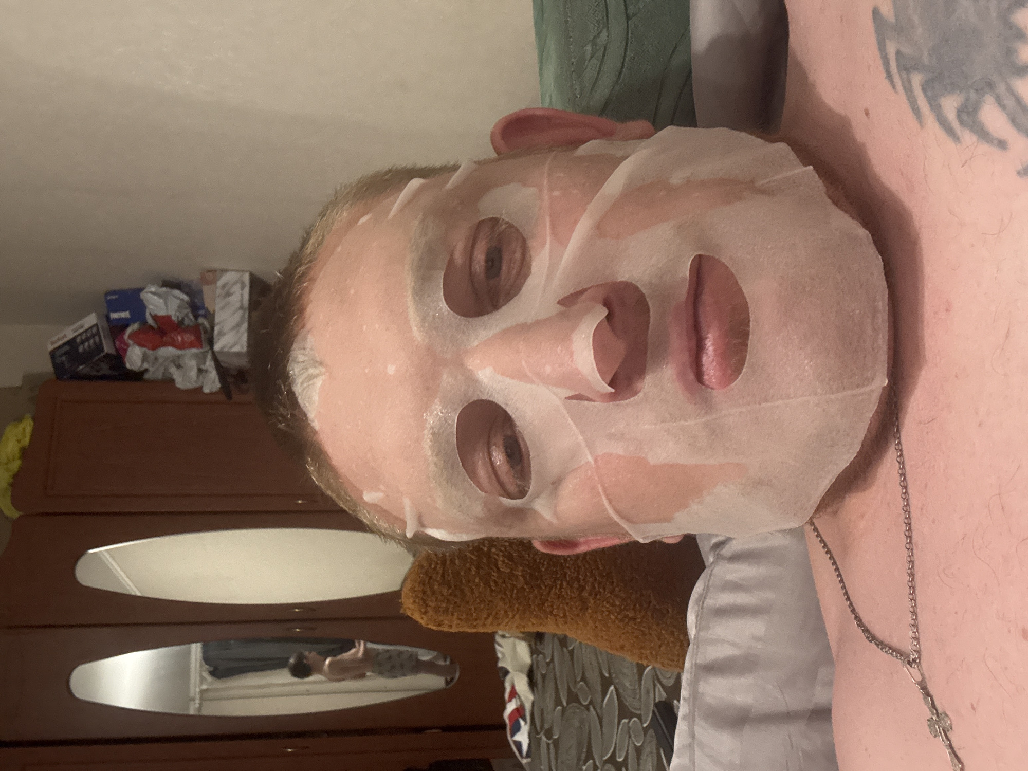A man, his face covered by a white sheet mask with eye and mouth openings, is lying in bed. He is wearing a silver chain necklace and has a tattoo on his chest. In the background, a closet with mirrors reflects a young boy standing in the room. The room appears to be a bedroom.