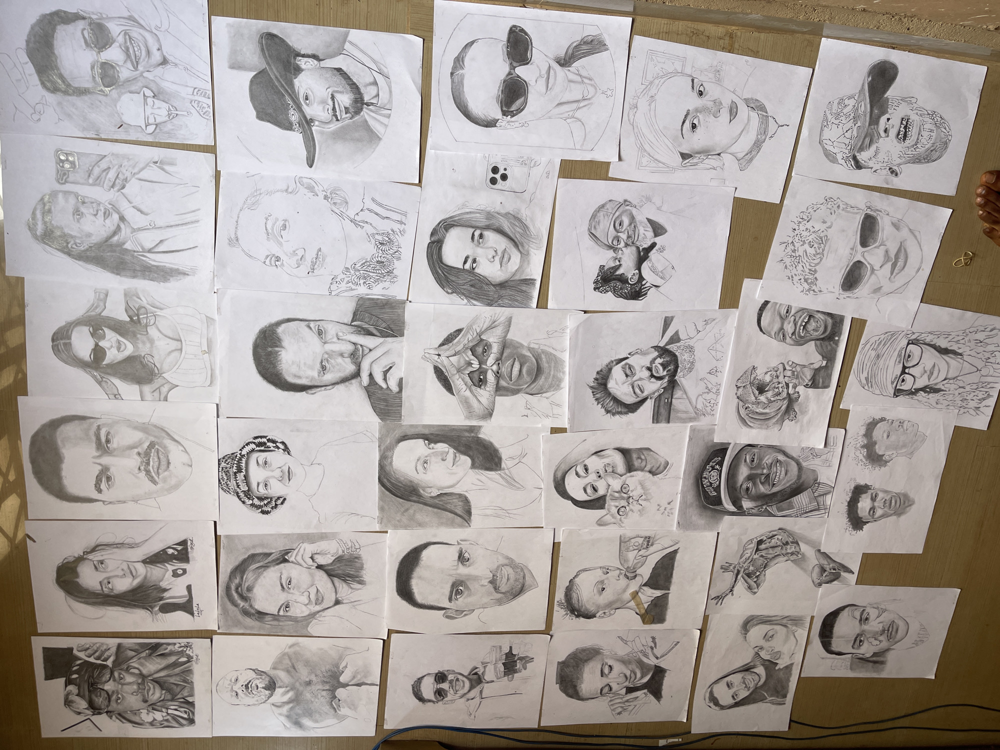The image shows a collection of roughly drawn pencil portraits spread out on a dull brown surface, likely a floor. The portraits depict various individuals, some wearing sunglasses, hats, and other accessories. A hand or foot is partially visible in the upper right corner of the image. A small gold-colored metal ring lies next to the foot. A blue or black electrical cable is barely visible at the bottom edge of the image. Given the context, the drawings were likely created in Jos, Nigeria.