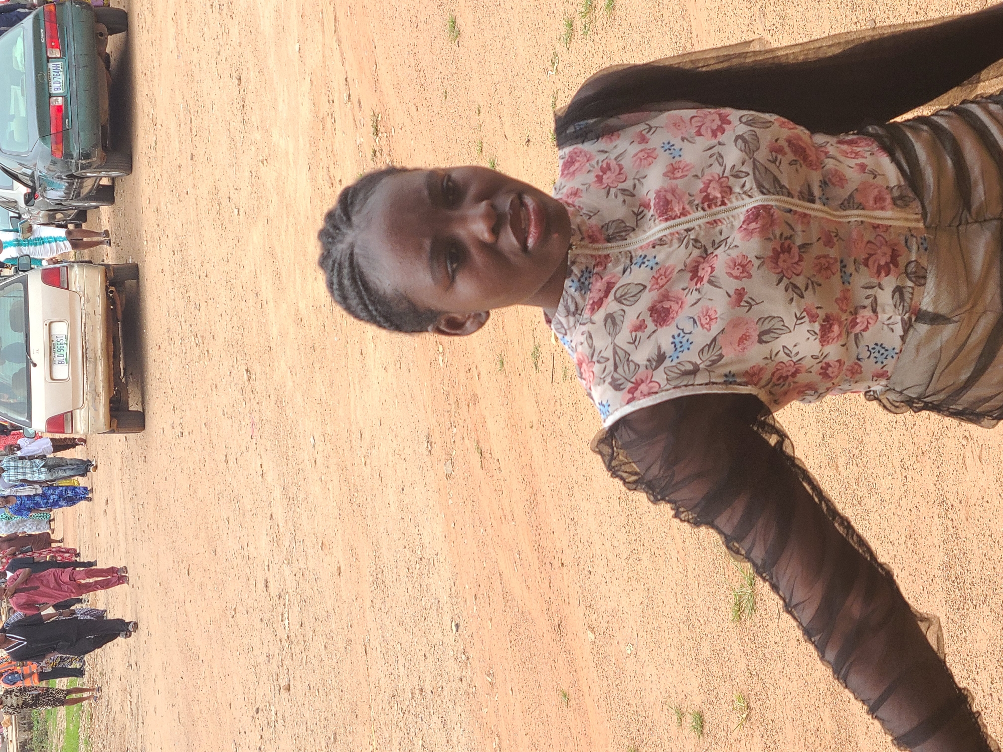 A young woman is standing on a dirt ground in Gura Top, Nigeria. She is facing the camera. She wears a floral top with a zipper and a black see-through shawl. Behind her, a group of people are standing in a line near two cars.
