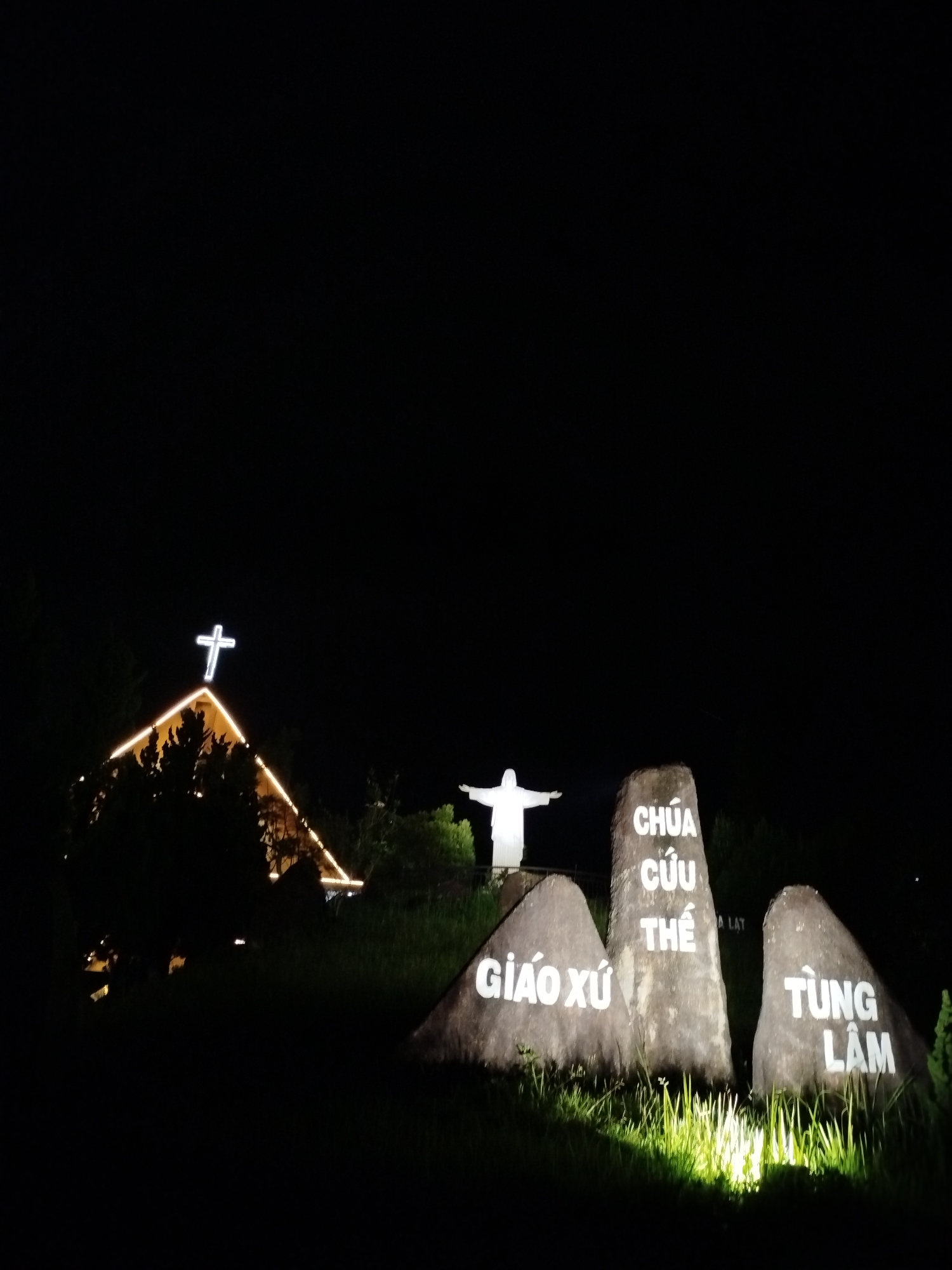 A nighttime view in Lạc Dương, Vietnam shows a church with a lit cross on its roof, a statue of Jesus with outstretched arms, and three large stones inscribed with Vietnamese text. The text on the stones reads "GIÁO XỨ" on the left stone, "CHÚA CỨU THẾ" on the center stone, and "TÙNG LÂM" on the right stone. All elements are illuminated against a dark sky.
