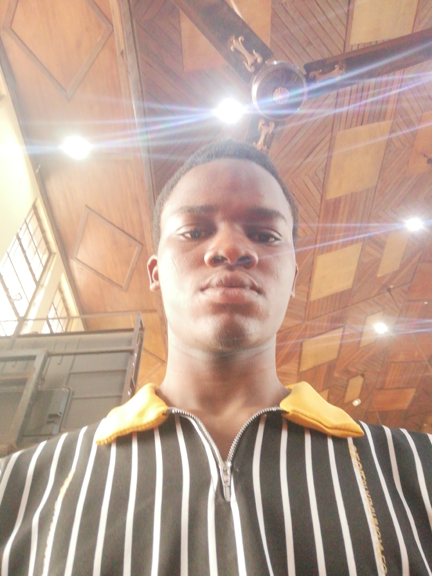 A medium shot of a dark-skinned man wearing a black and white striped shirt with a yellow collar. He appears to be indoors. Above him is a decorative wooden ceiling, with a ceiling fan and recessed lights. To the left are windows and a metal structure. The location is plausibly Enugu, Nigeria, based on the individual's appearance and general environment, but no definitive landmarks are present.