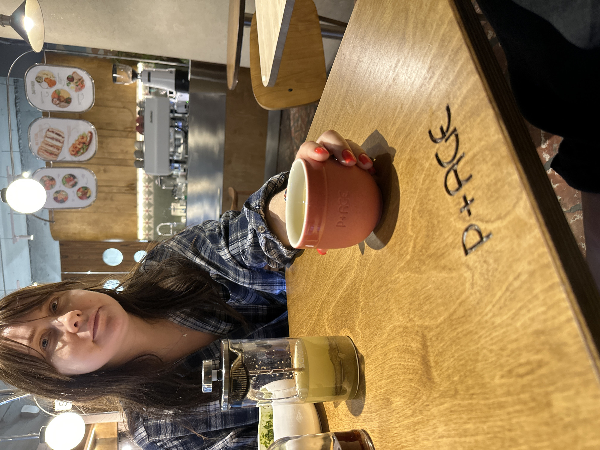 The image shows a woman sitting at a wooden table in a cafe, likely in Sochi, Russia, given the name "D+RGE" etched into the table. She is wearing a blue and black plaid shirt and holding an orange mug with red nail polish on her fingers. On the table, there's a French press filled with a green beverage, a white bowl, and another small glass container. The cafe's interior is visible in the background, with menu boards displaying food items and a coffee machine. The lighting suggests it is daytime.