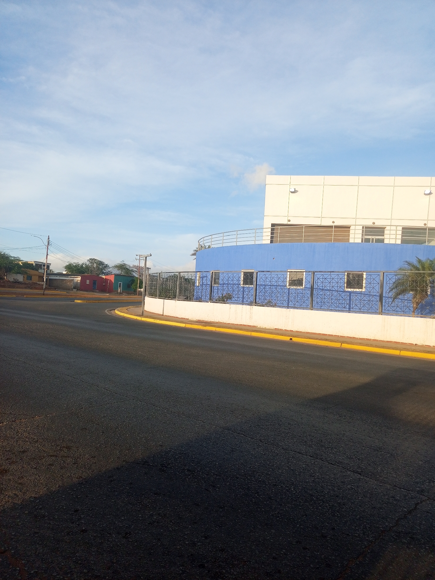 The image shows a street corner in Juan Griego, Venezuela. A two-story building with a white upper level and a blue lower level stands at the corner. The lower level has a fence with a geometric pattern and square openings. A palm tree is visible behind the fence. A yellow curb lines the edge of the road. In the background, there are small, colorful buildings, including one that is red and one that is green. Utility poles and power lines are also visible. The road surface is dark asphalt, with sunlight casting shadows. The sky is blue with scattered clouds.