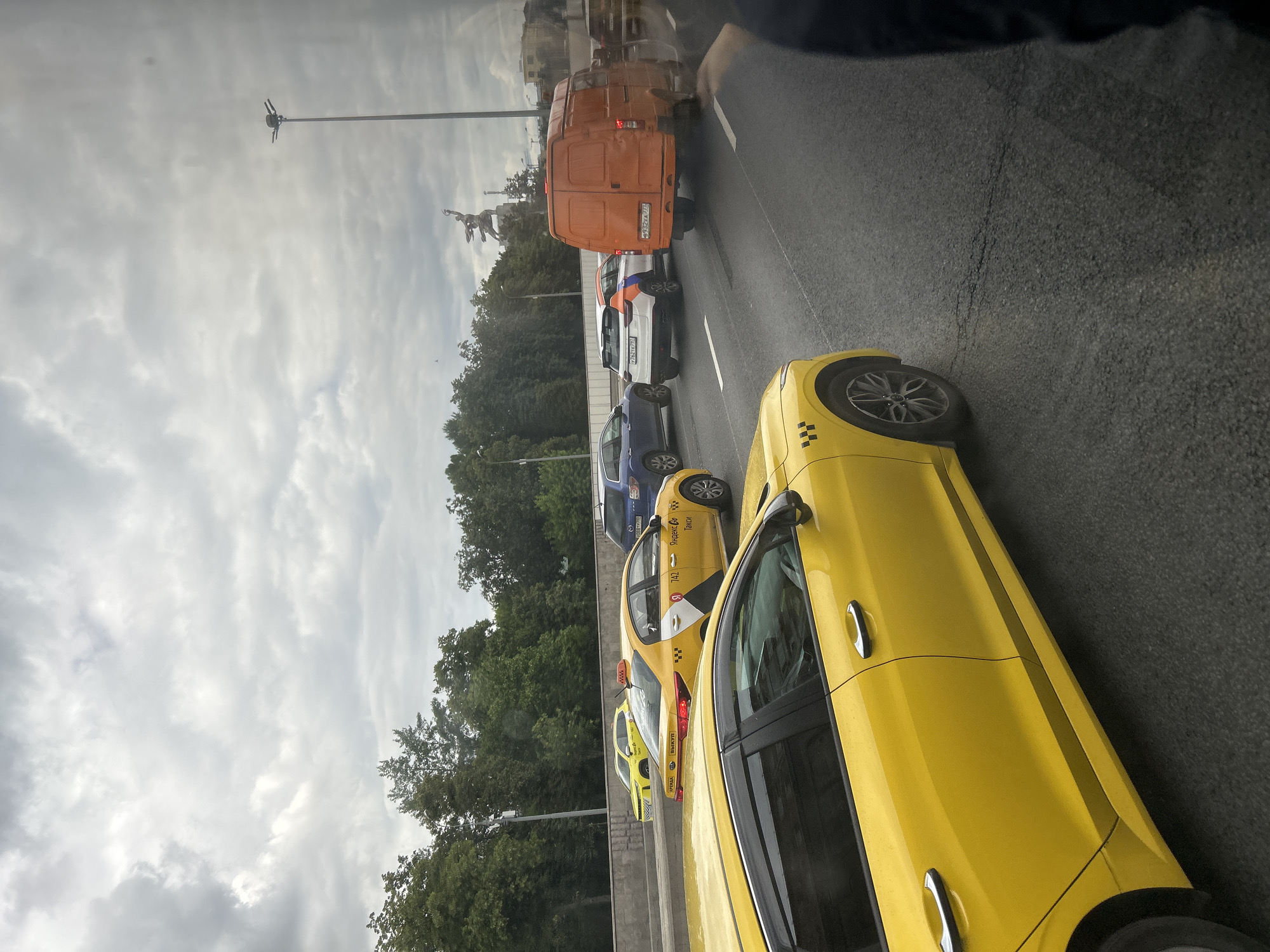 The image shows a traffic jam on a cloudy day in Moscow, Russia. Several cars are visible on a multi-lane road, including yellow taxis, a white and orange SUV, and an orange van. In the background, trees line the road, and a statue is visible in the distance, likely a landmark of the city. The sky is overcast with gray clouds. The foreground is dominated by the side of a yellow car.