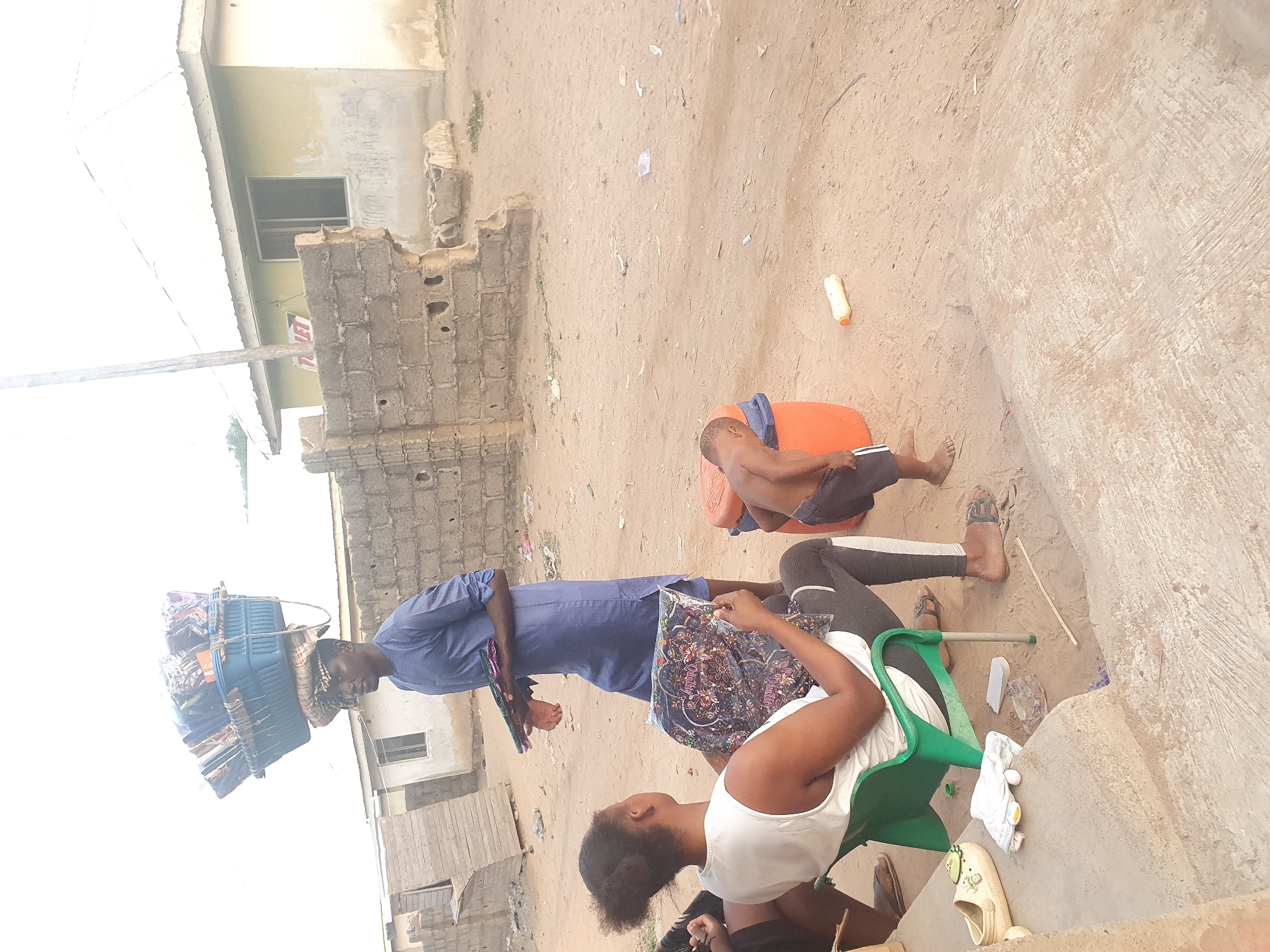 A man in Lokoja, Nigeria, is carrying a large bundle of goods balanced on his head. He is wearing a blue collared shirt. He stands near two seated people and appears to be presenting them with packaged textiles. One person is seated on a green plastic chair, wearing a white tank top. The other person is seated on an orange plastic bucket. They are barefoot. The scene is outdoors, with sandy ground and simple buildings visible in the background.