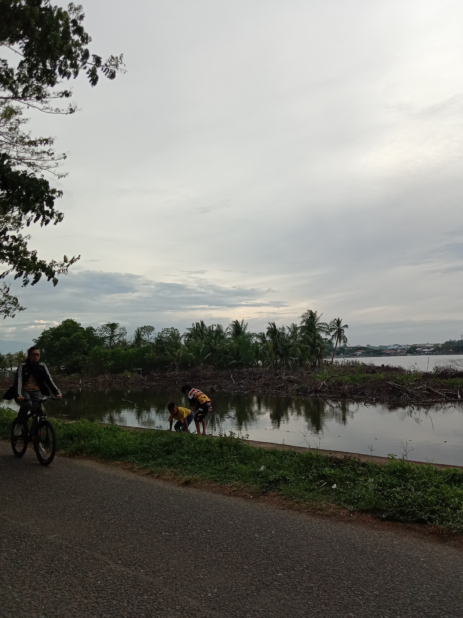 The image captures a scene in Lhokseumawe, Indonesia. A man rides a bicycle on an asphalt road. Two children are near the water's edge, possibly interacting with the water. The water area shows signs of debris, possibly driftwood or remnants of trees. Trees and vegetation are visible across the water. The sky is overcast.