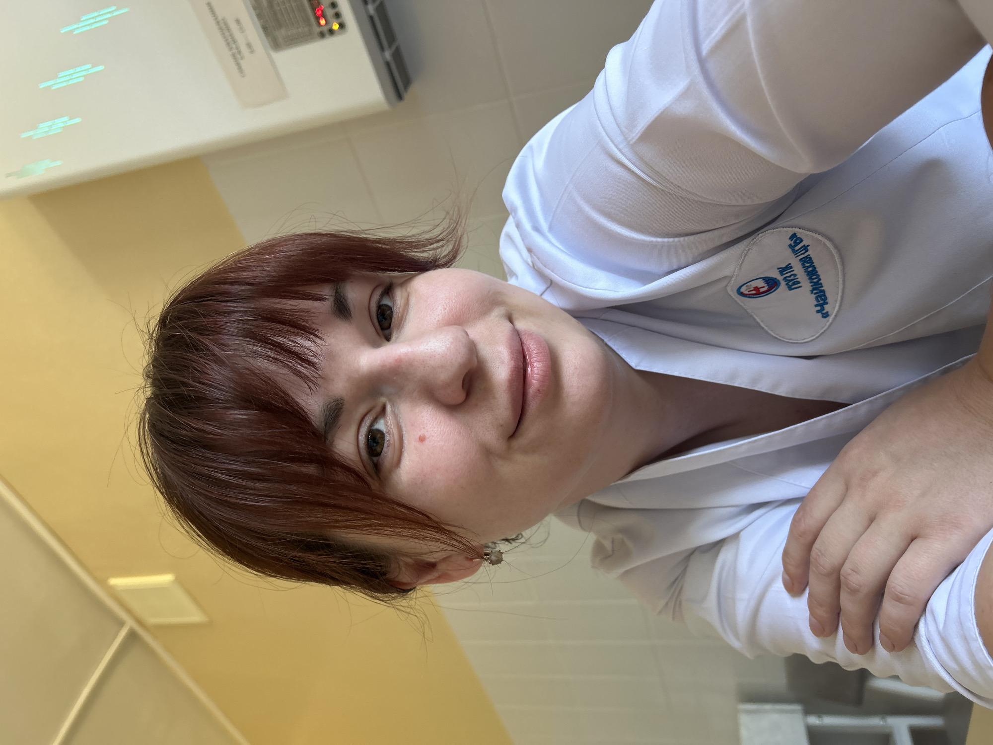 A woman with reddish-brown hair is wearing a white medical coat with a patch that reads "ГБУЗ ПК Чусовская ЦРБ" on it. The location is in the city of Фоки, Russia based on the text on the coat. She is looking directly at the camera with a slight smile. The background includes a cream-colored wall and a piece of equipment with buttons and a display. She's either in a medical facility or clinic. Her hand is visible in the lower right corner of the frame.