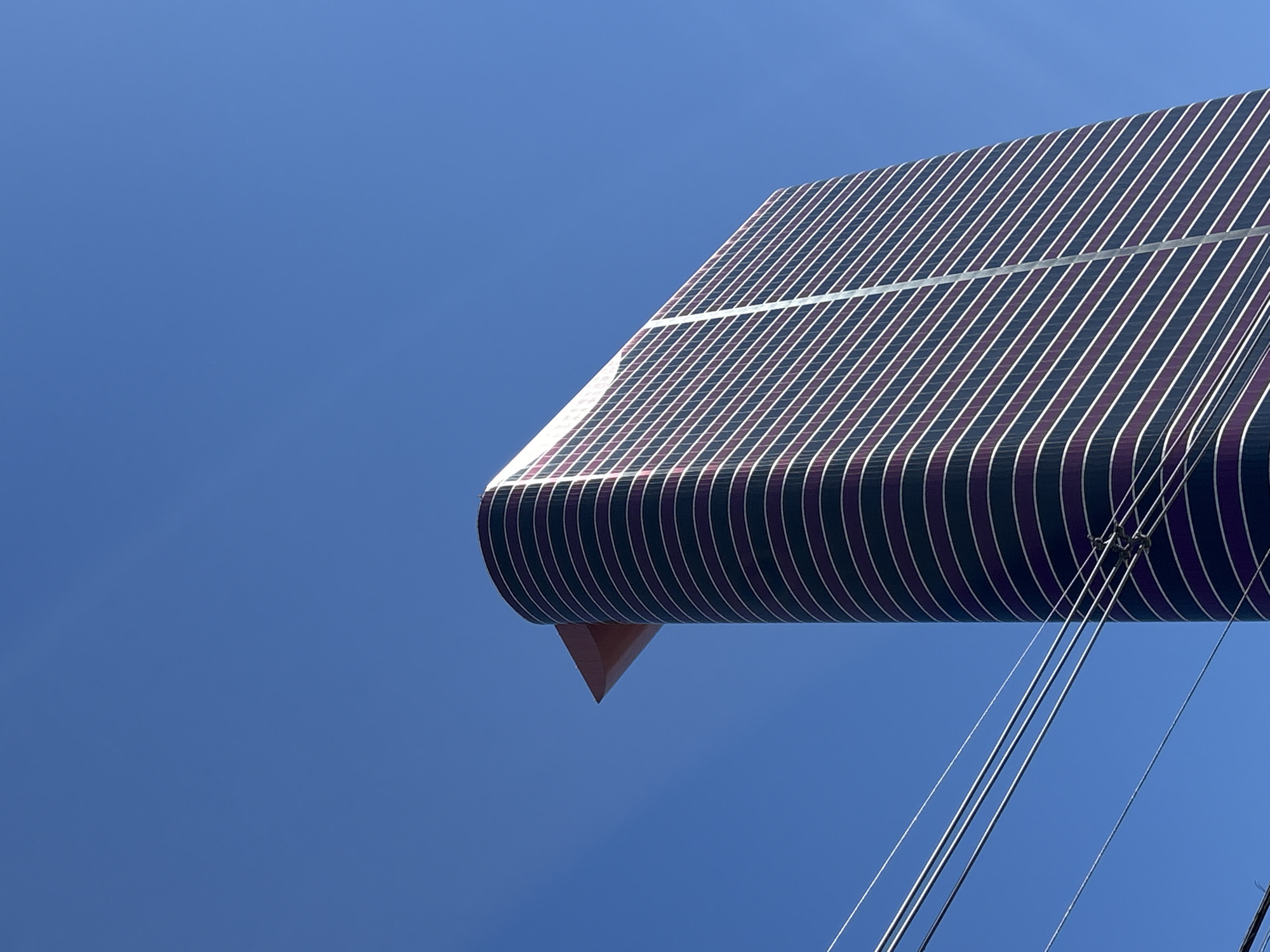 A modern, curvilinear skyscraper dominates the frame, its dark purple and white striped facade reaching towards a clear, bright blue sky. The building's distinct architectural feature is a sharp, triangular protrusion at its upper section, rendered in a reddish-brown hue. In the lower left of the image, a series of power lines stretch horizontally across the scene, their dark cables contrasting with the vibrant sky and the sleek building. The perspective is from a low angle, looking up at the skyscraper, emphasizing its height and imposing presence. The scene appears to be set in a city, given the presence of the skyscraper and the utility lines. This building is the Transamerica Expo Center in São Paulo, Brazil.