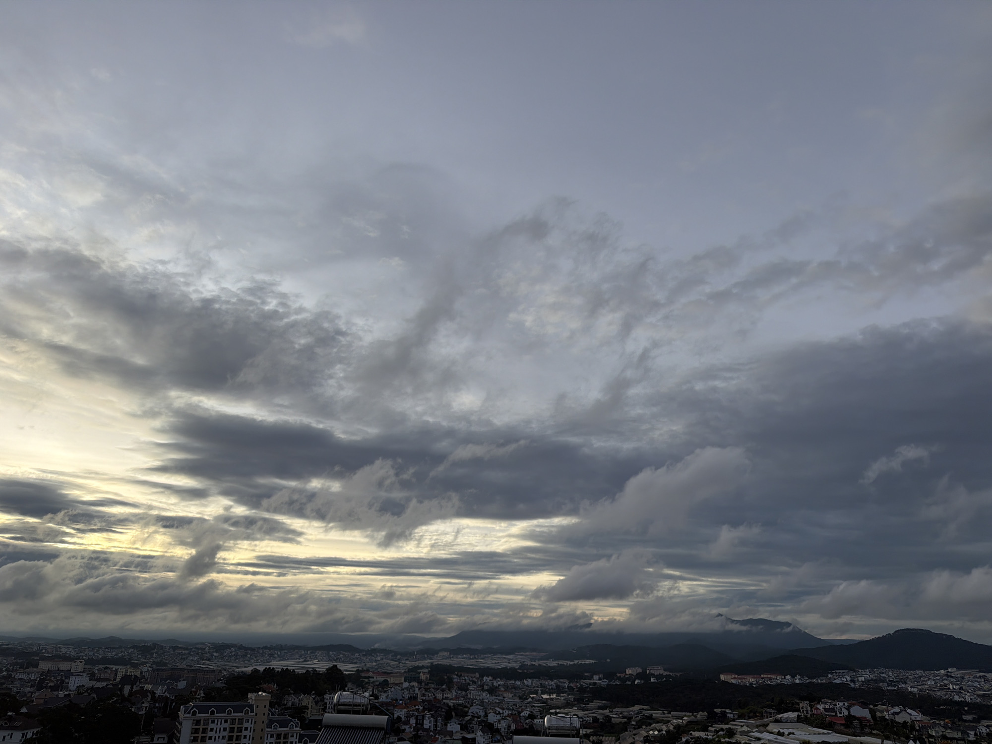 The image shows a panoramic view of a city, likely Xuân Trường - Đà Lạt, Vietnam, under a dramatic sky. The sky is filled with heavy, dark clouds that are being illuminated by a soft, diffused light, possibly from the rising or setting sun. Streaks of brighter light break through the clouds, creating a textured and dynamic atmosphere. Below the sky, a city sprawls across a hilly landscape. Buildings with varying architectural styles are visible, including residential areas and some larger structures. In the foreground, solar water heaters are present on some rooftops. The horizon is lined with dark, rolling hills and mountains, partially obscured by the clouds and mist. The overall impression is one of a serene yet moody landscape, captured during twilight hours.