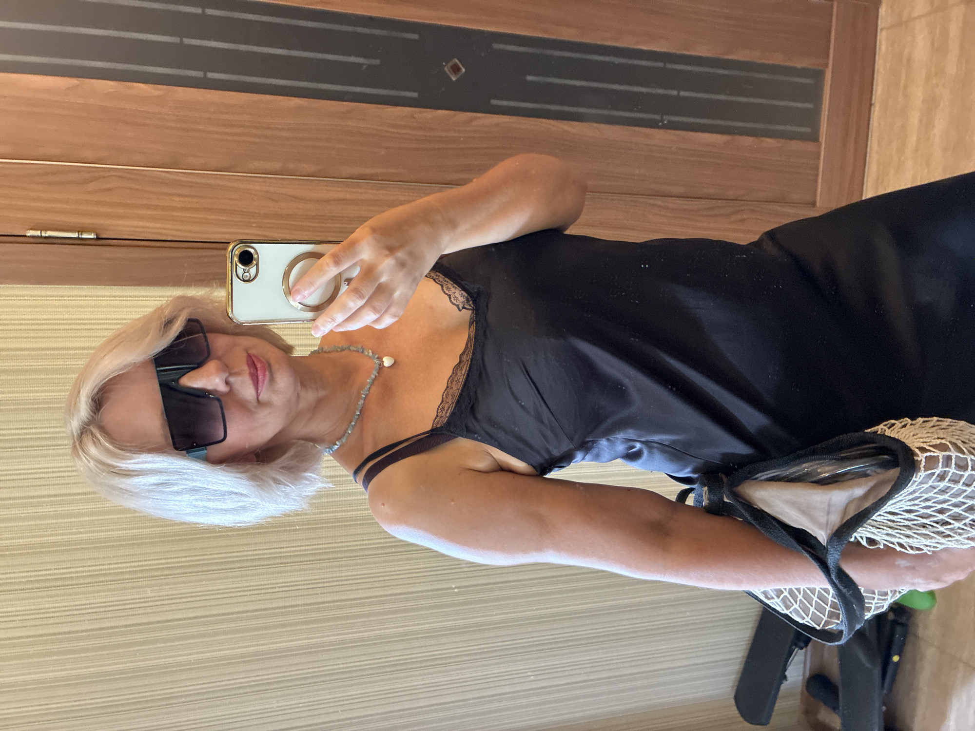 In an indoor setting, a blonde woman with short hair, wearing large sunglasses and a black slip dress with lace trim, takes a selfie in a mirror. She is also wearing a beaded necklace with a heart-shaped pendant and is holding a white mesh bag with a black trim in her left arm. Behind her, a wooden door with vertical glass panes is visible. The floor appears to be tiled.