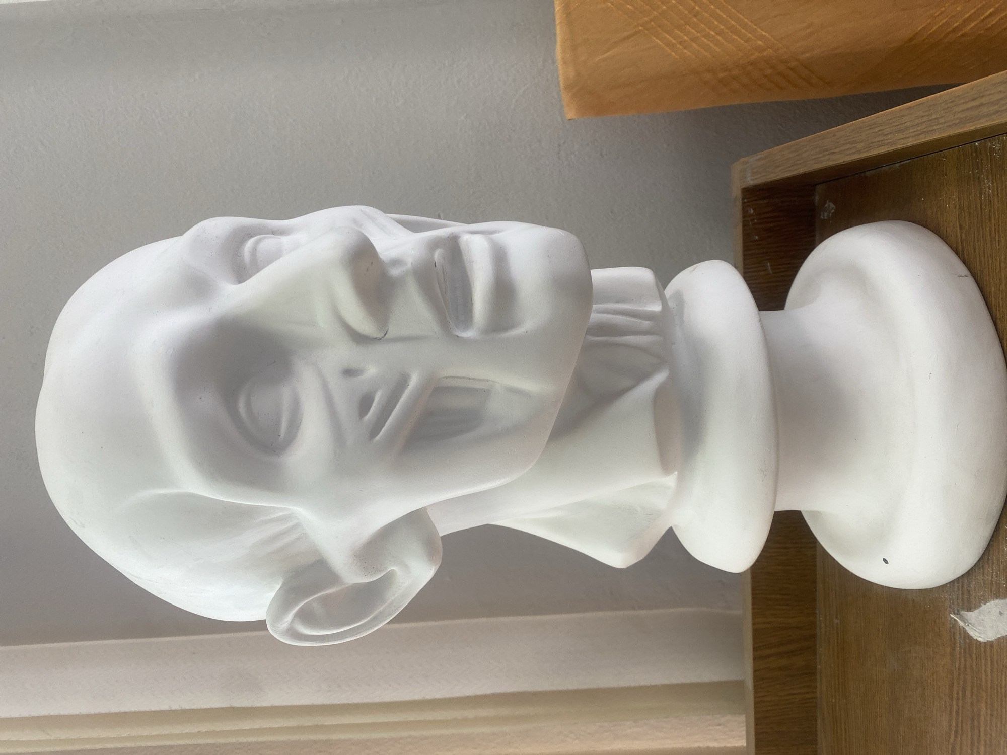 A white plaster bust of a classical figure rests on a wooden surface. The bust is of a human head and neck. The head is inclined slightly to the right. The wooden surface appears to be a shelf or tabletop. The background includes a white wall. There is no indication of the image's location being in Yekaterinburg, Russia.