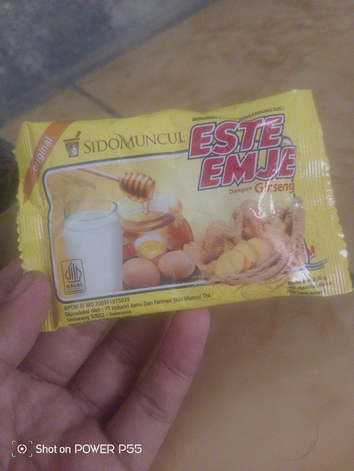 A person's hand is holding a yellow packet of "SIDOMUNCUL ESTE EMJE DENGAN GINSENG". The packet features an illustration of a glass of milk, eggs, honey, and ginseng. Text on the packet indicates it is a beverage containing milk and ginseng. There is also a Halal certification logo. Below the illustration, there is text that reads "BPOM RI MD 200911015039 Diproduksi oleh : PT Industri Jamu Dan Farmasi Sido Muncul Tbk Semarang 50552 - Indonesia". The image was taken in Lebaksiu, Indonesia, and the watermark "Shot on POWER P55" is visible in the bottom left corner. The background appears to be a tiled surface and a wooden surface.