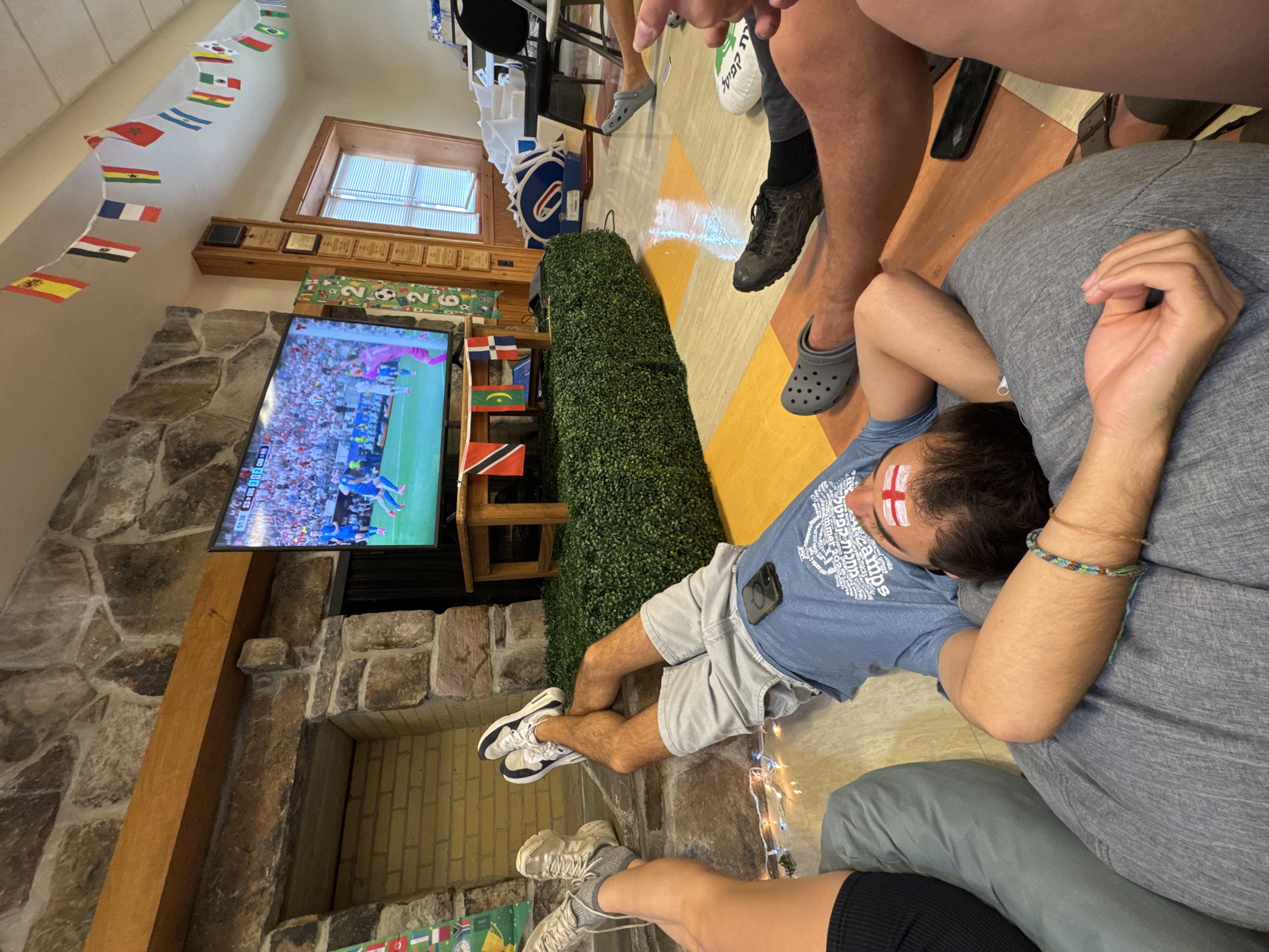 In Washington Township, United States of America, a group of young adults are gathered in a room, likely a common area or recreational space, to watch a soccer match on television. The room features a prominent stone fireplace with a wooden mantel. A large flat-screen television is mounted above a wooden table, which is covered with artificial green turf. The television displays a soccer game with a stadium filled with spectators.
Several individuals are seen lounging comfortably, with one person lying on their back on a large gray beanbag chair, looking up at the TV. This person has a bandage or face paint, possibly representing the English flag, on their forehead. Another person is lying on the floor with their legs crossed, wearing shorts and a blue t-shirt, with a phone placed on their stomach. Two other individuals' legs are visible in the foreground, one wearing athletic shoes and socks, and another wearing Crocs. The floor is a mosaic of yellow and orange tiles.
The room is decorated with a string of small flags, likely representing various countries participating in a tournament. A wooden cabinet with multiple plaques is mounted on the wall to the right of the television. In the background, other furniture and objects, including chairs and what appear to be large cardboard cutouts, are visible. String lights are draped near the floor, adding a festive atmosphere. The overall scene depicts a relaxed and engaged gathering of friends enjoying a sporting event.